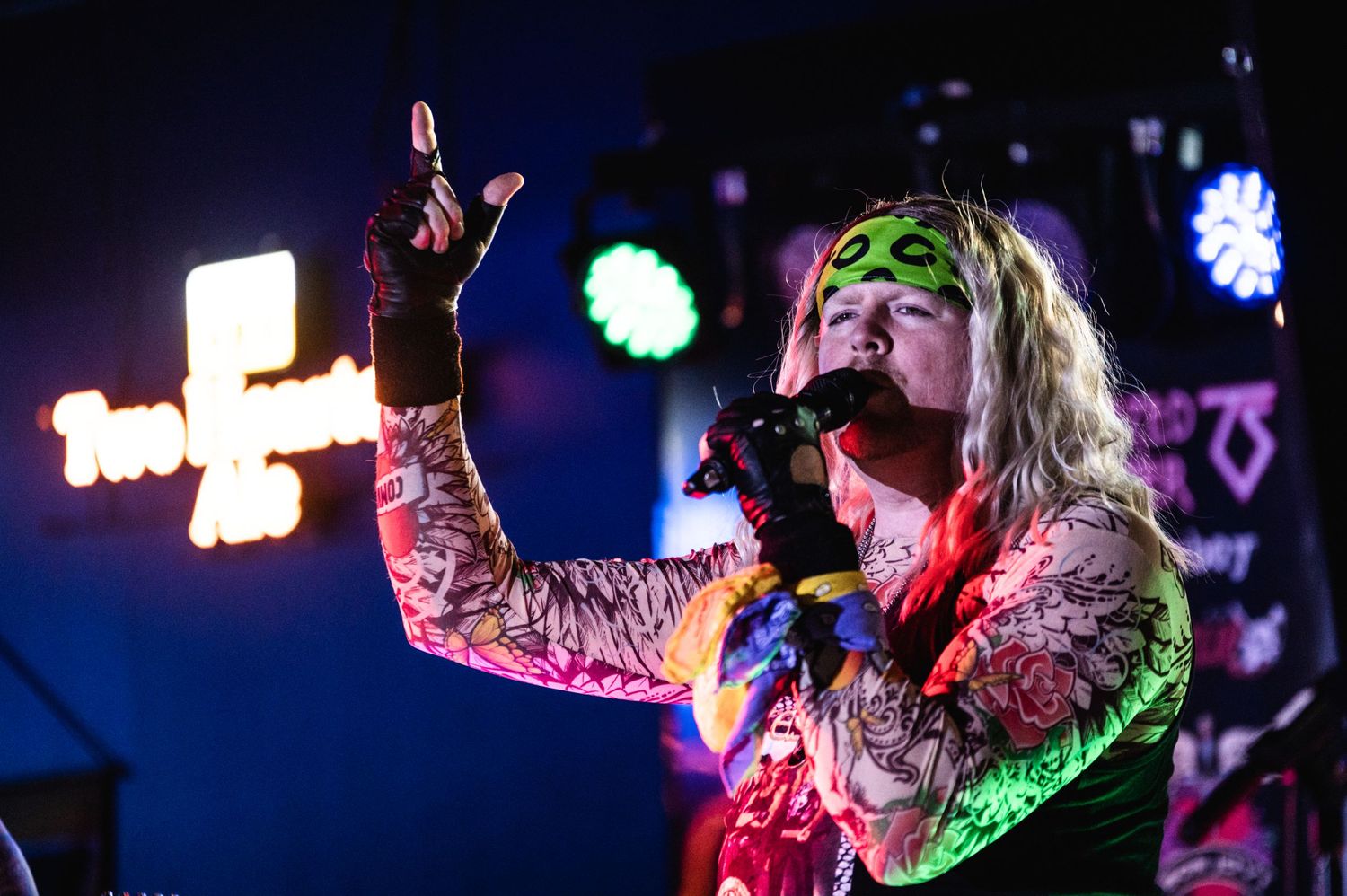 Concert performer in stage lighting singing with arms raised wearing bright green face paint.