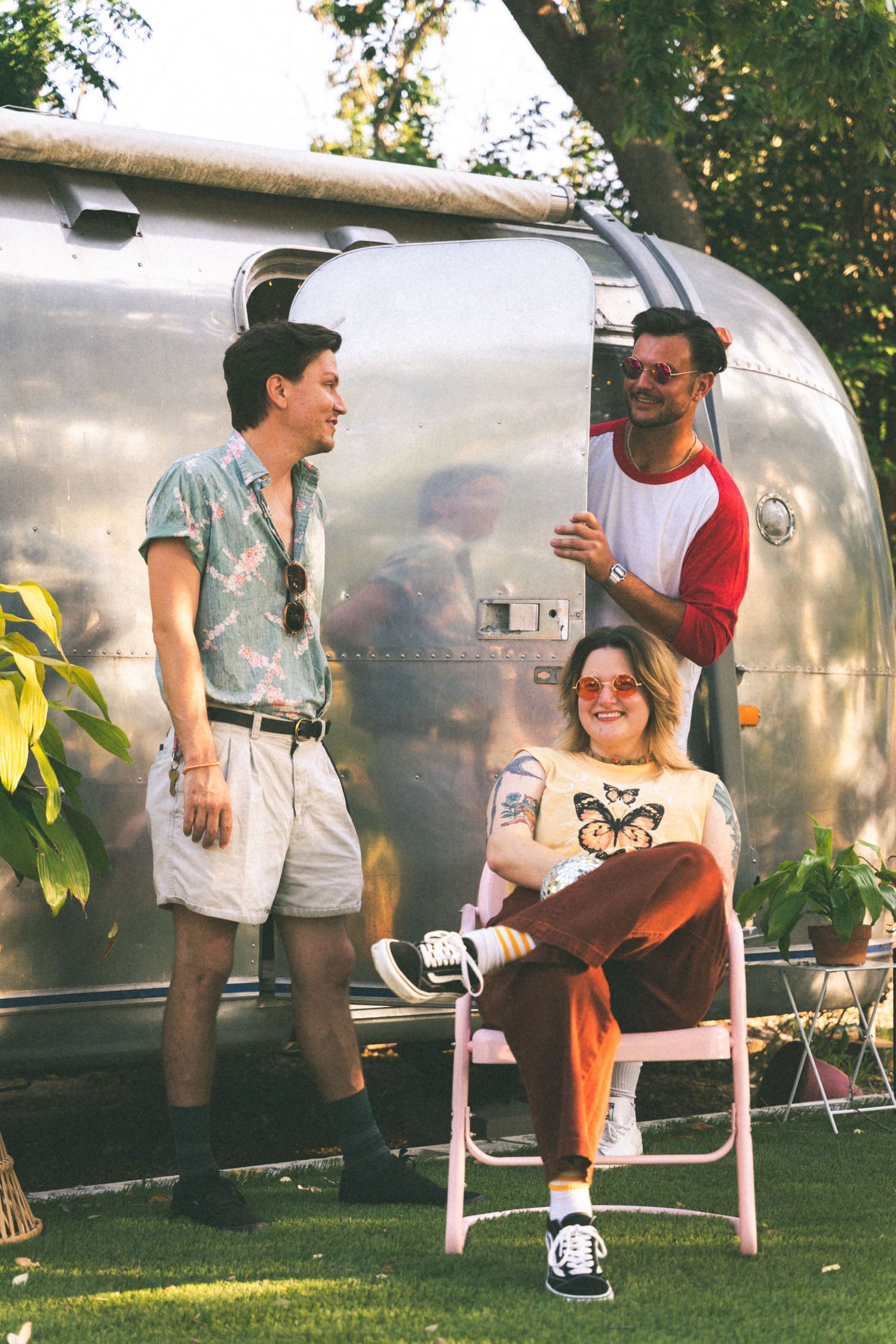 Three friends hanging out by a silver Airstream trailer on a sunny day.