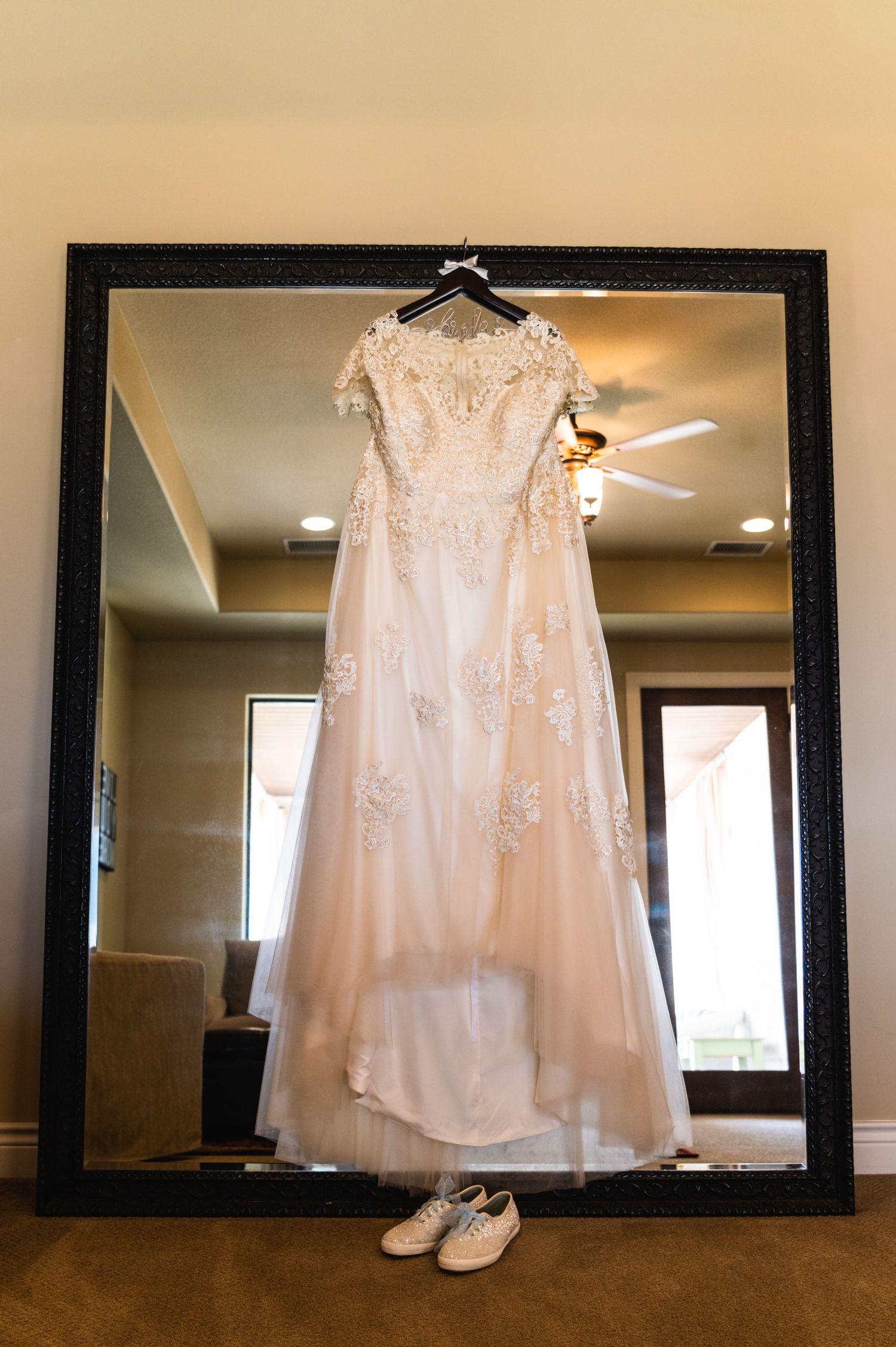 A beautiful white lace wedding dress hangs elegantly on a hanger against a large mirror with white shoes beneath.