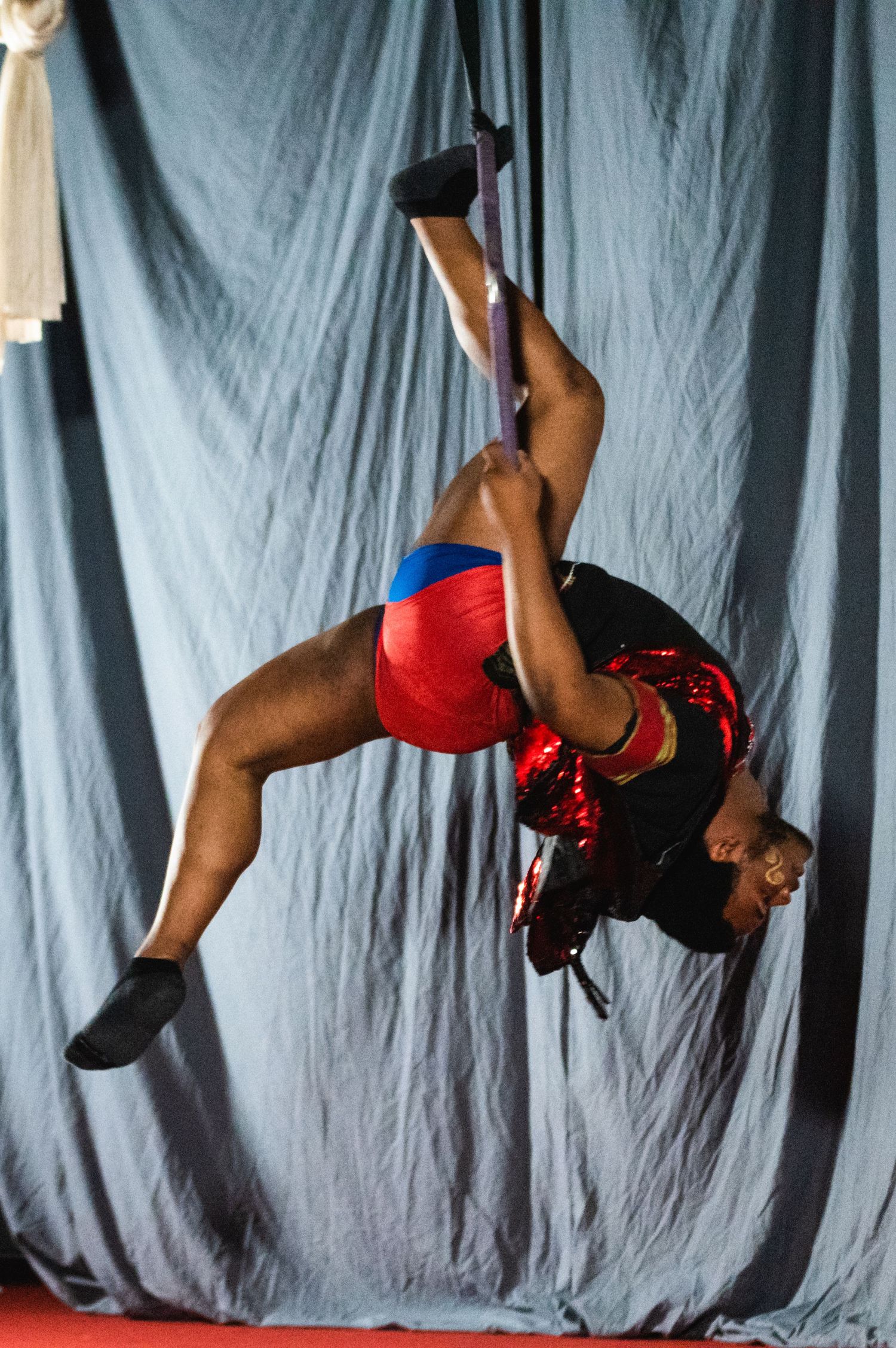 Aerial performer executes dynamic pose in red costume against grey backdrop.