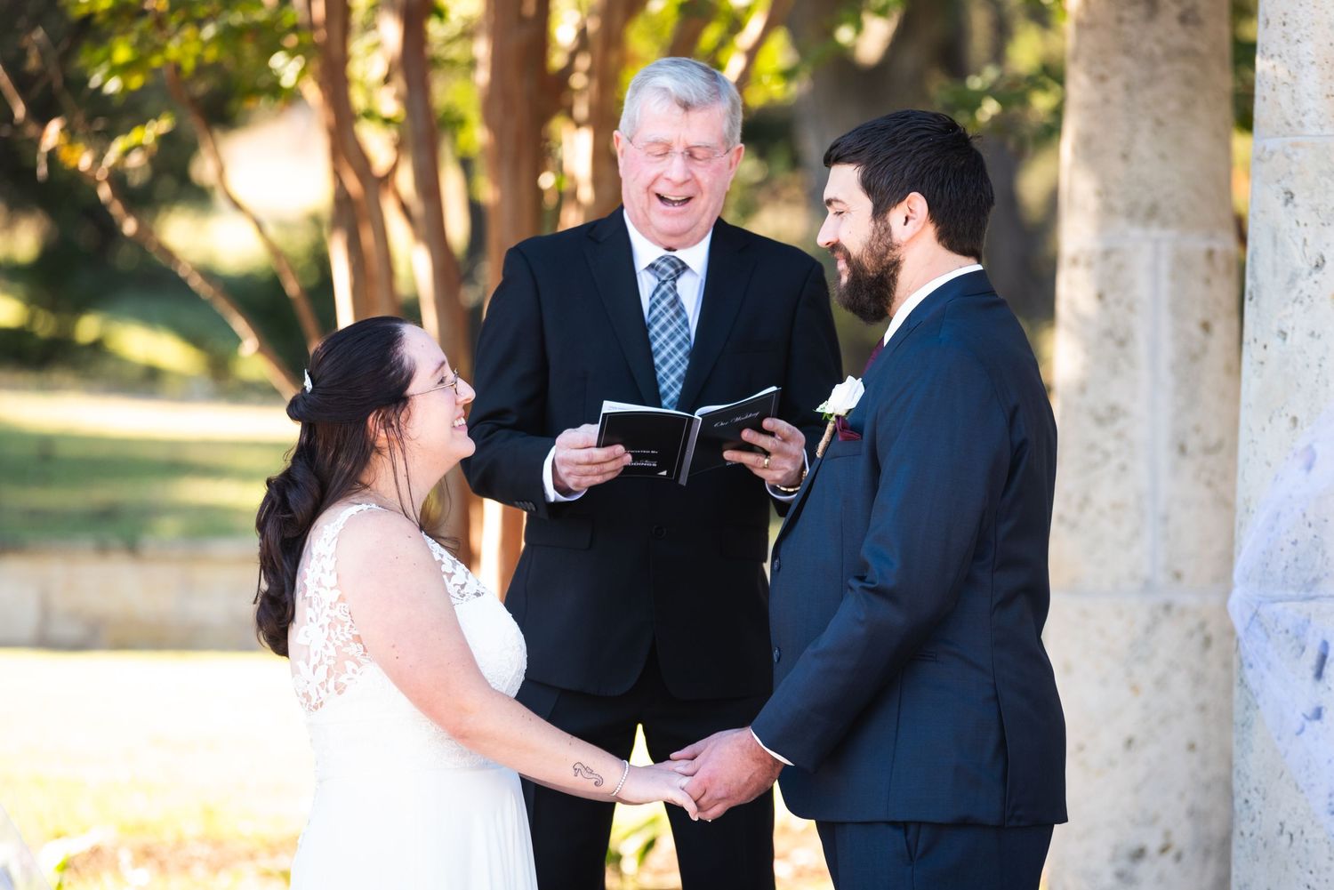 Outdoor wedding ceremony with couple and officiant standing under pillared structure.