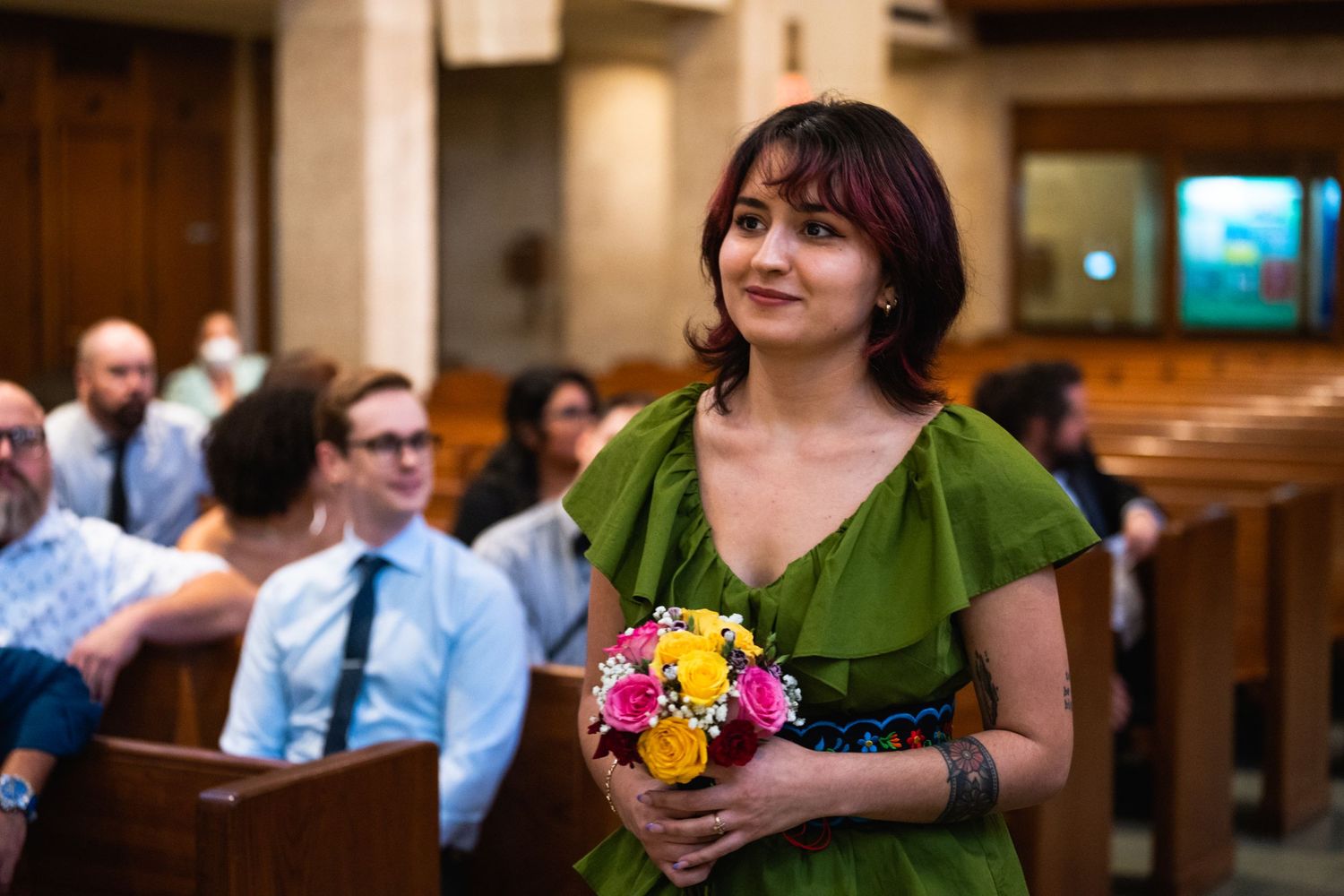 Guest in green dress holds colorful flower bouquet at church ceremony.