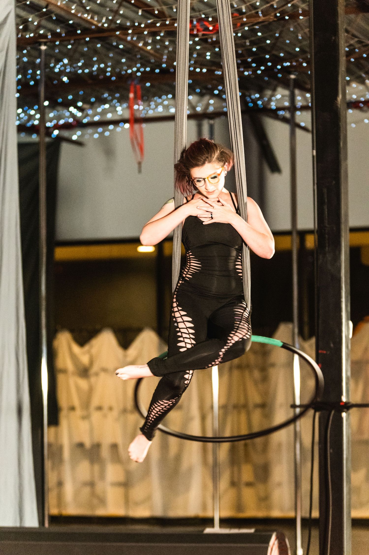 Aerial performer in black costume suspended gracefully from silk ribbons under twinkling lights.