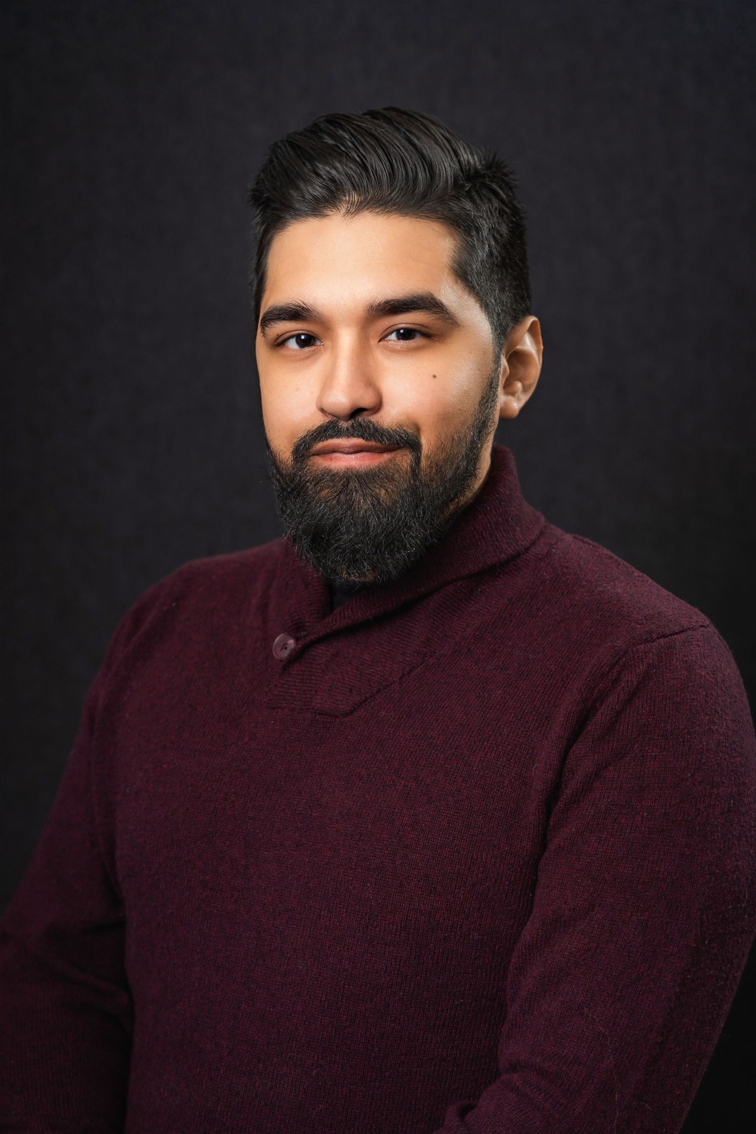 Professional headshot of person with beard wearing burgundy sweater against dark background.
