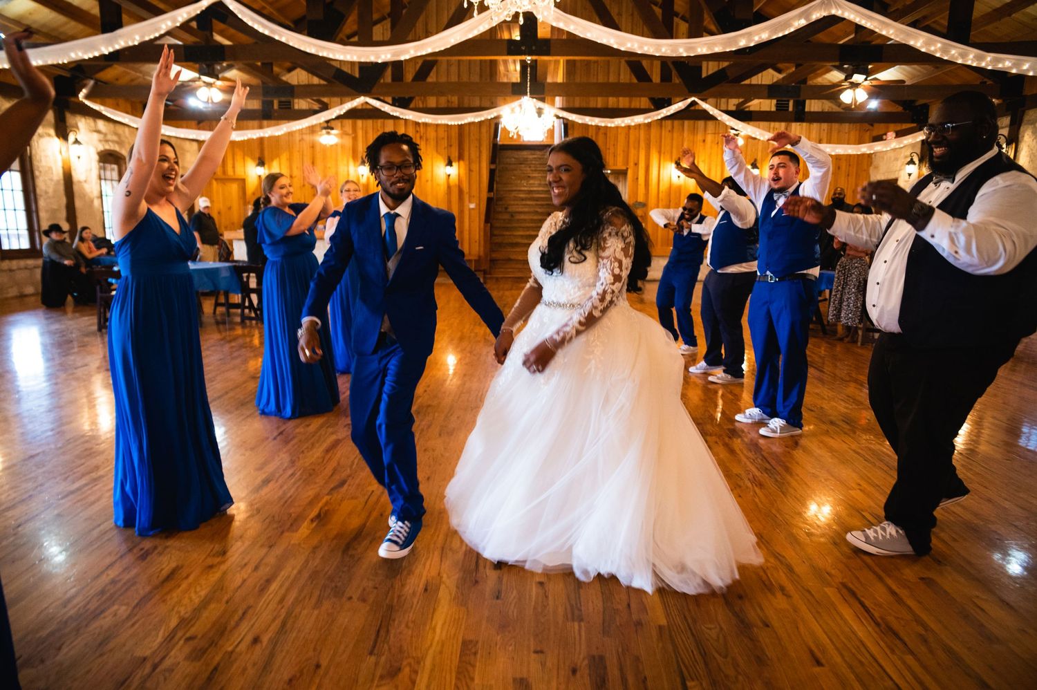 Wedding party dances in celebration on wooden floor with blue attire and white decorations overhead.