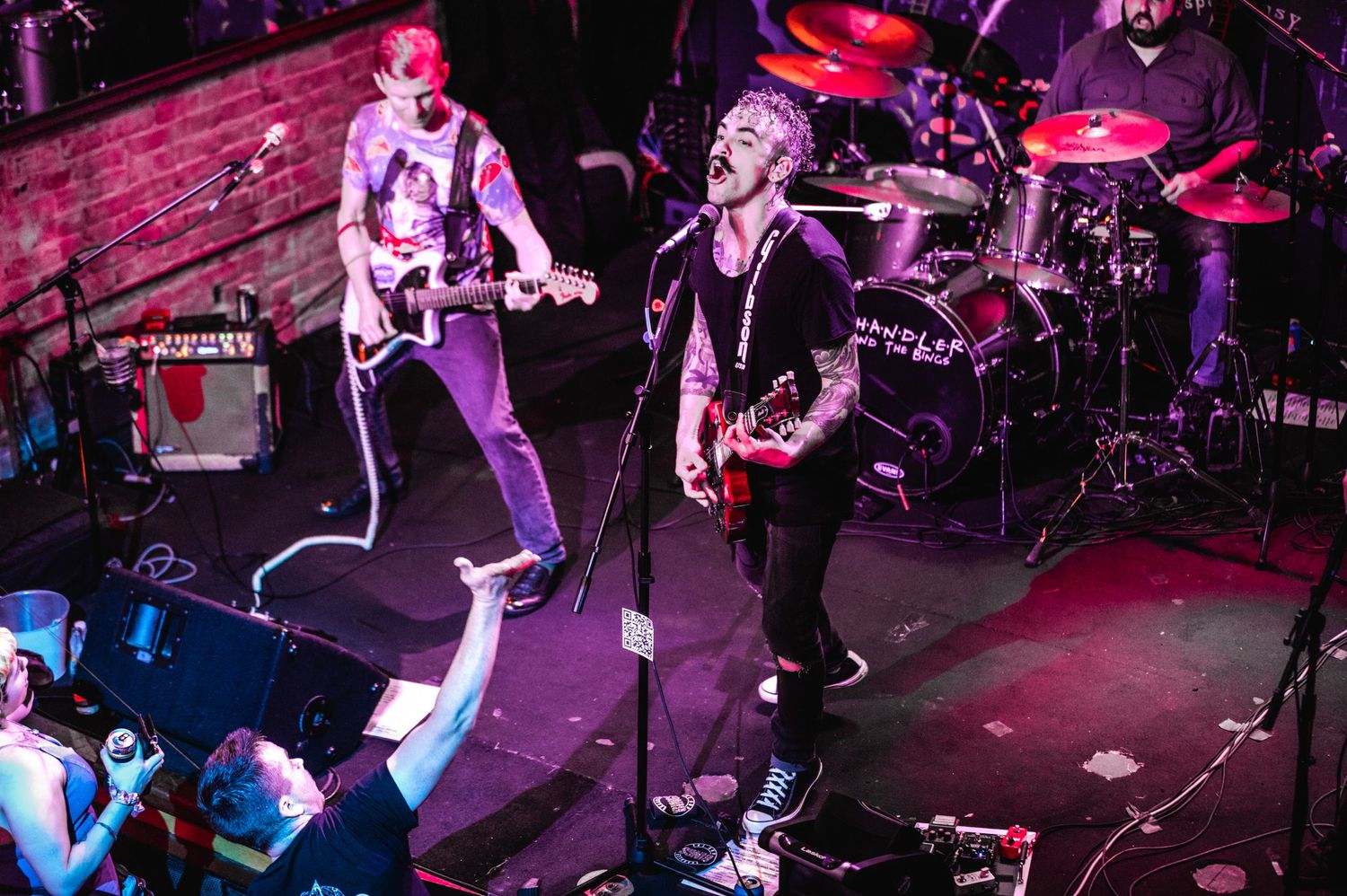 Musicians energetically performing on stage with crowd visible in foreground.