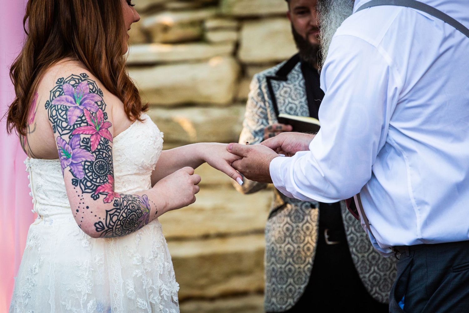 Close up of ring exchange during wedding ceremony with stone wall and pink decor.