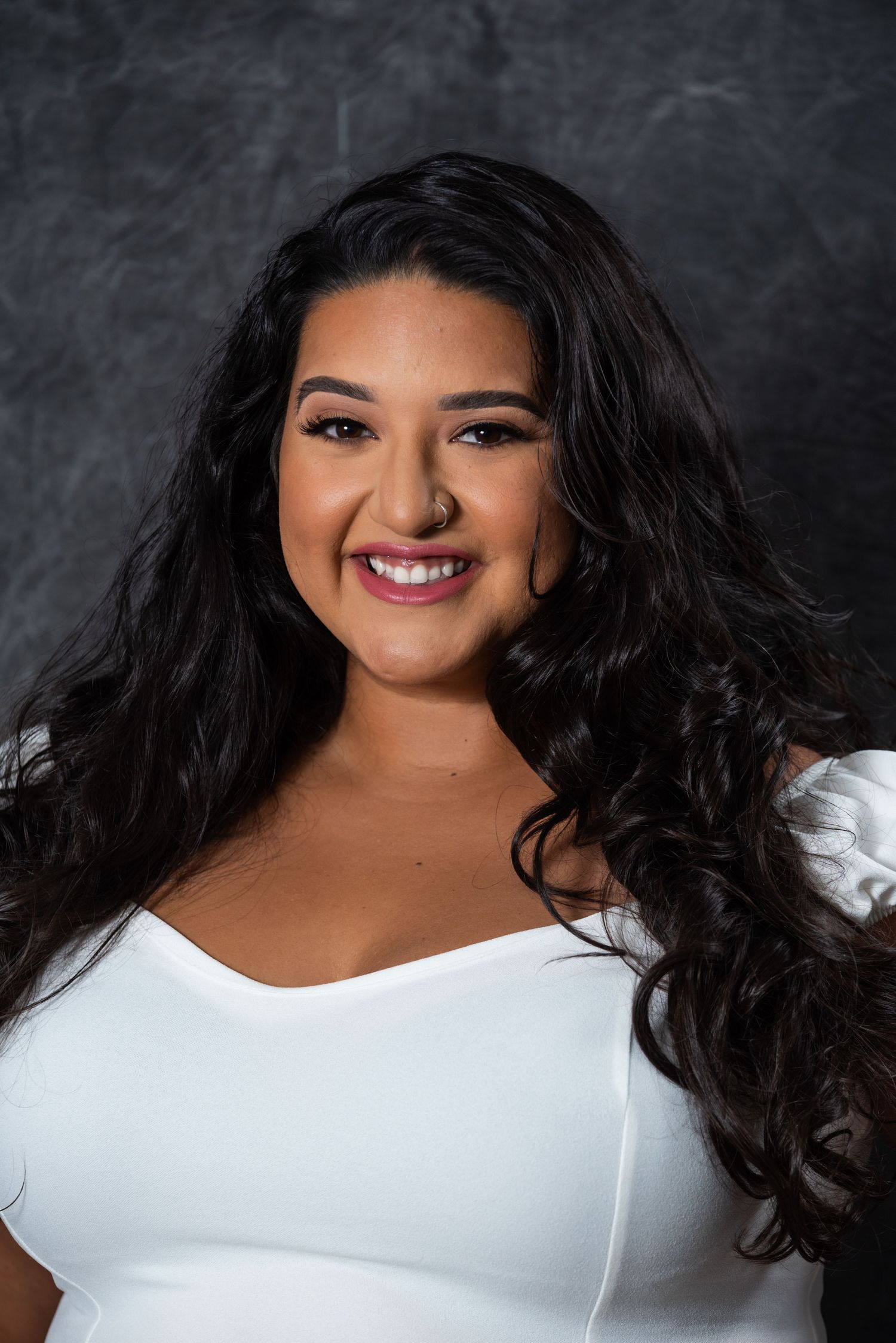 Studio headshot featuring flowing dark hair and a bright smile against a textured grey backdrop.
