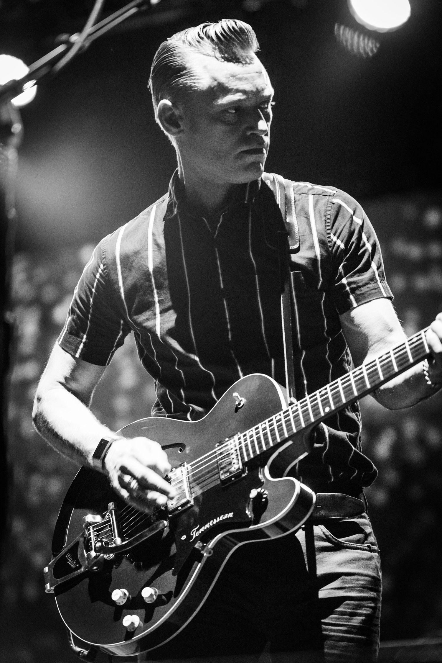 A musician plays a black hollow body guitar while performing on stage under dramatic lighting.