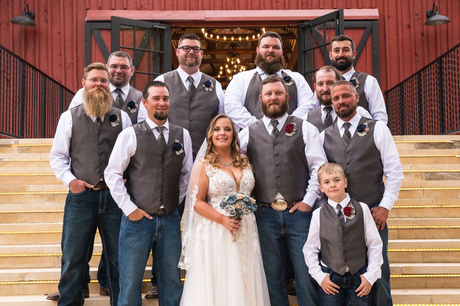 Wedding party stands on rustic barn steps wearing coordinating gray vests and casual attire.