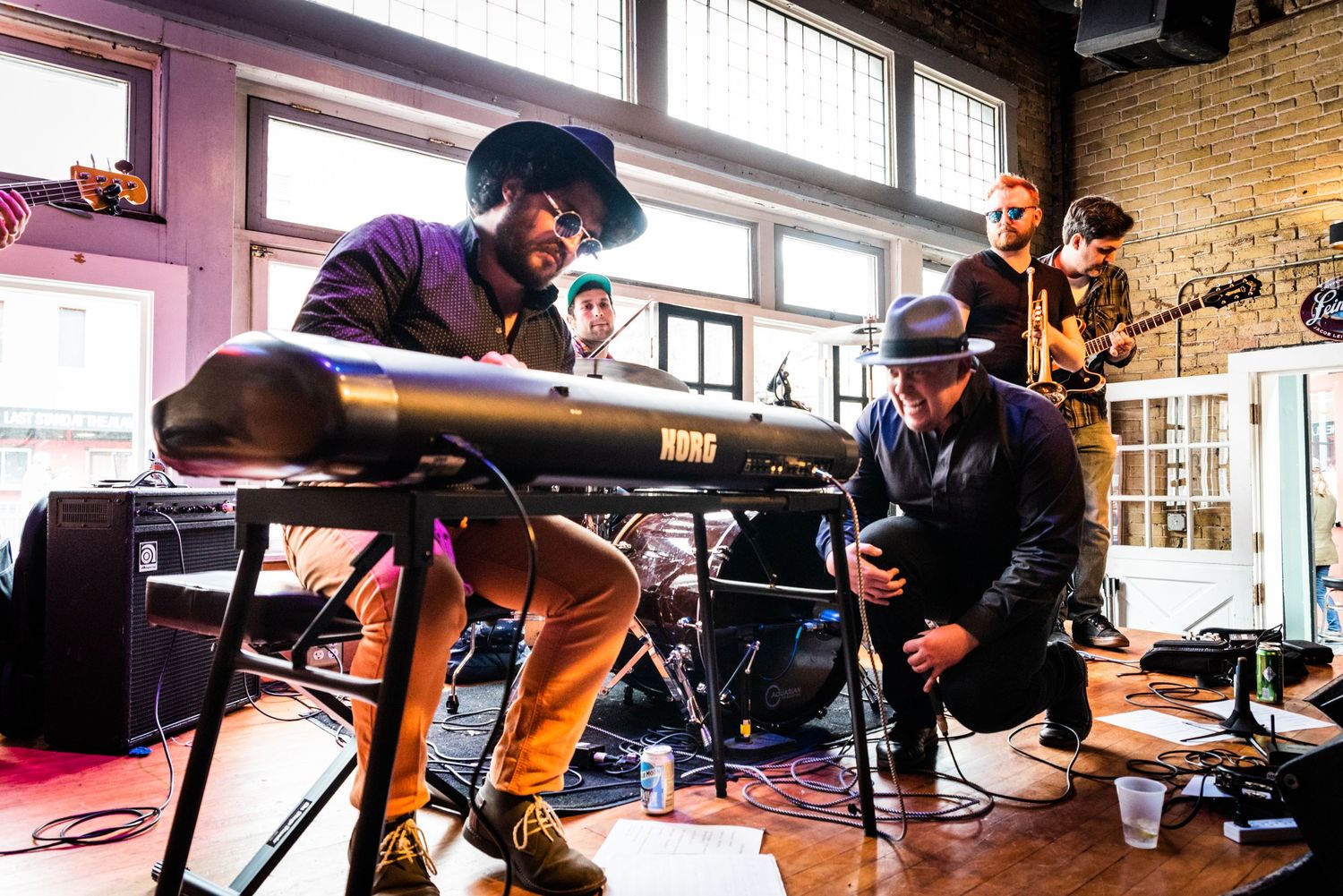 Musicians performing with keyboard setup in sunny venue with exposed brick interior.