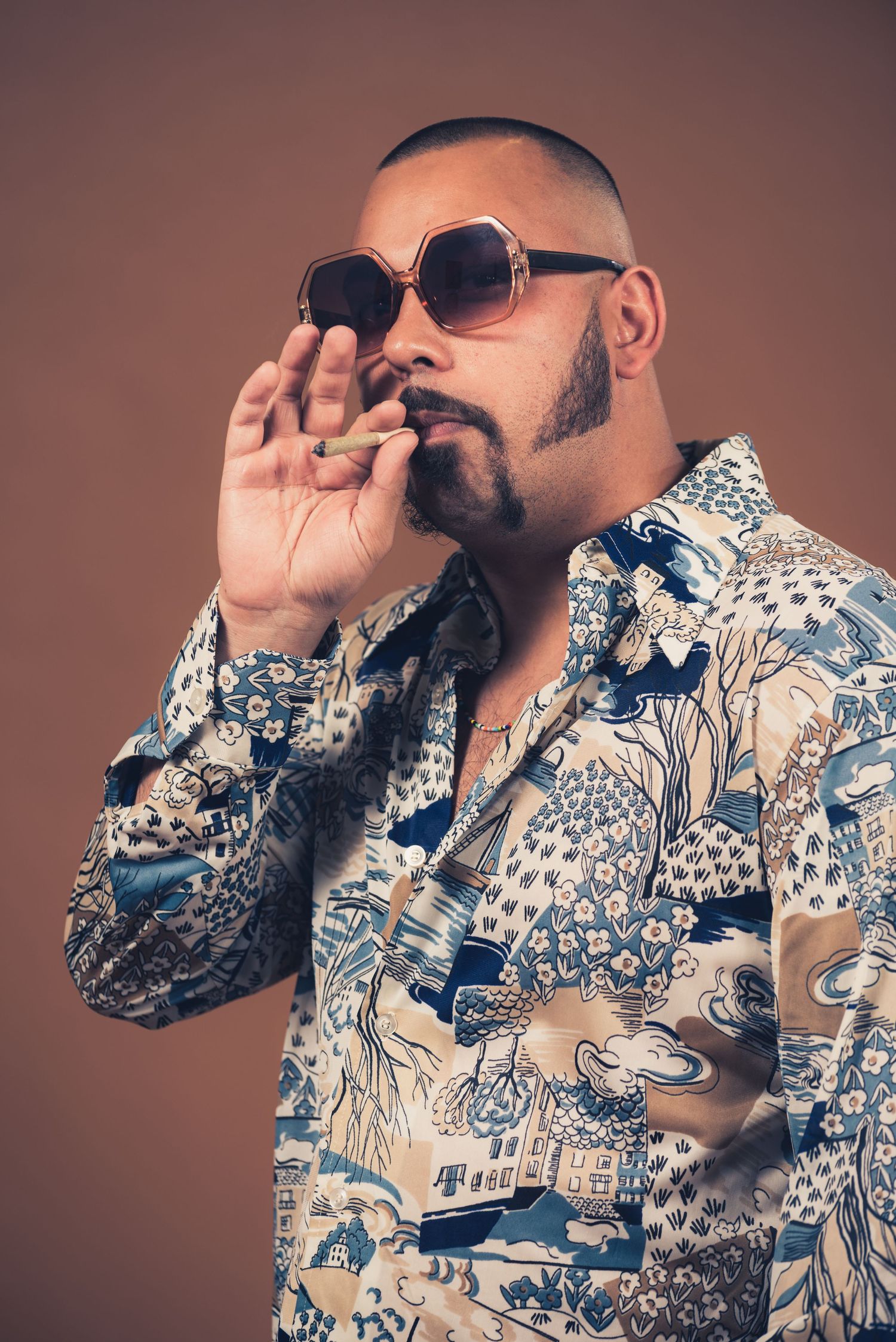 Person in patterned shirt and sunglasses posing with smoke effect against brown backdrop.