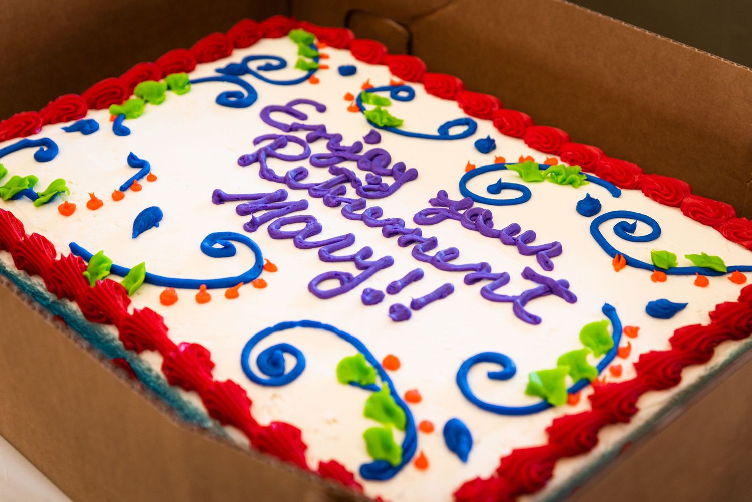 Decorated sheet cake with colorful swirls and festive writing in a cardboard box.