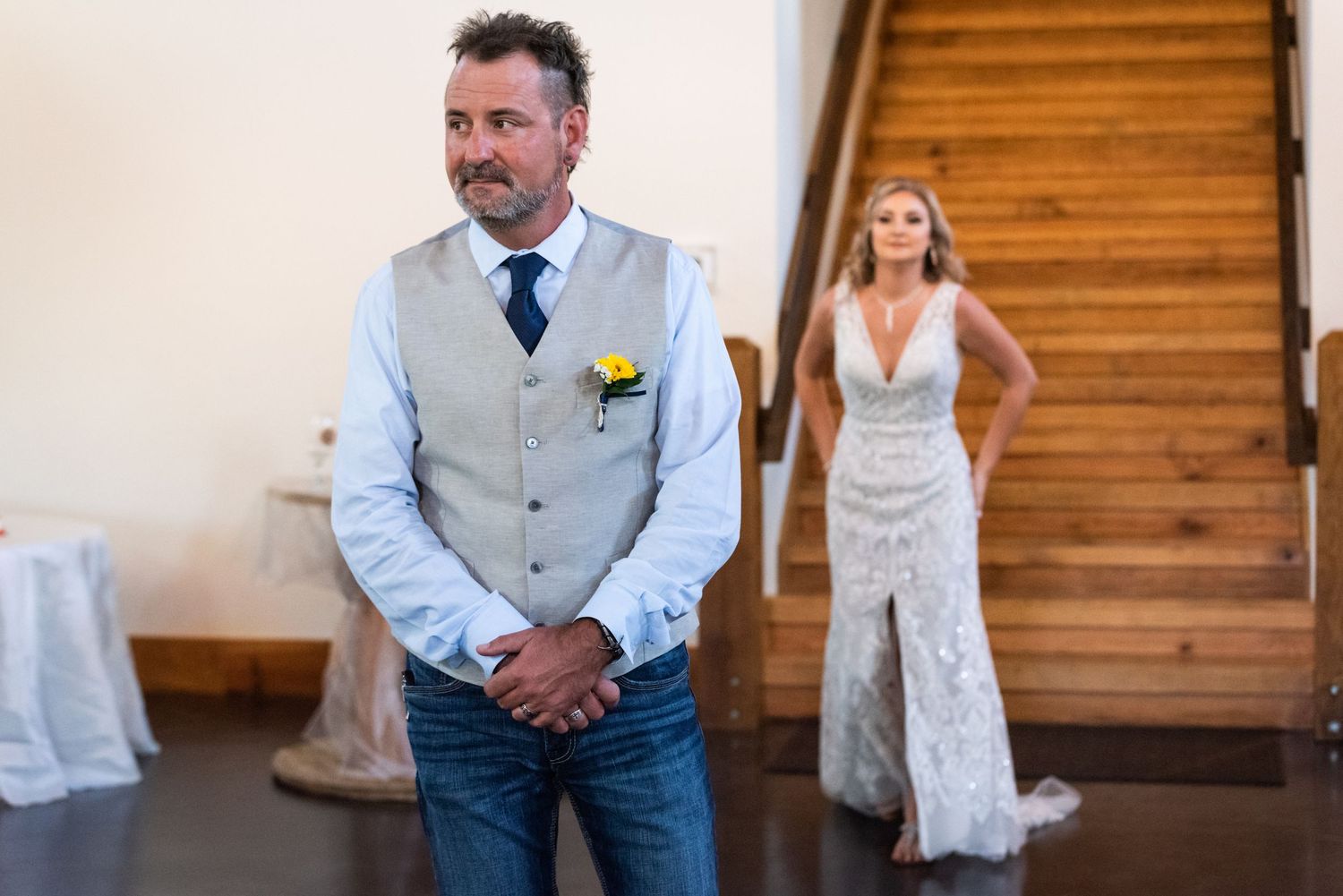 A touching wedding reveal scene with a groom in a light vest and jeans near wooden stairs.