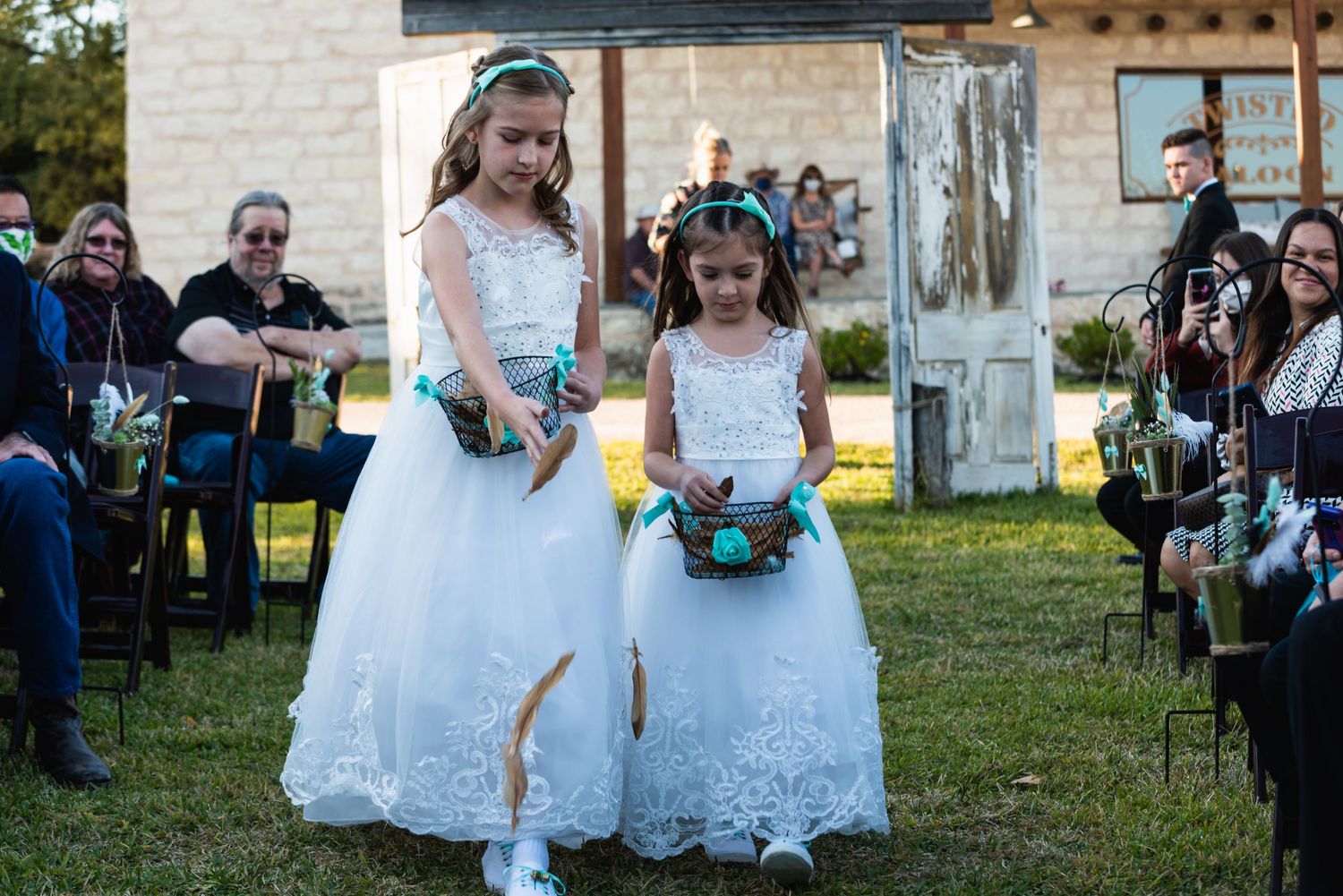 Two young flower girls in white lace dresses walk down outdoor wedding aisle with turquoise accessories.