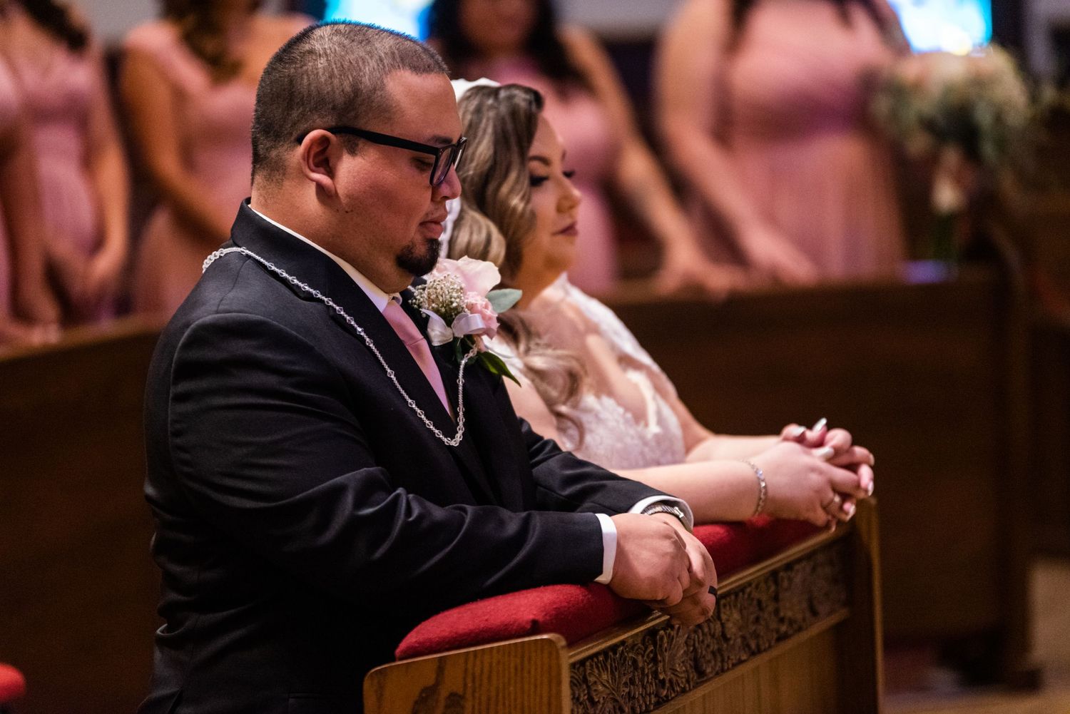 A person kneels in prayer during a church ceremony with pink-dressed attendees visible.