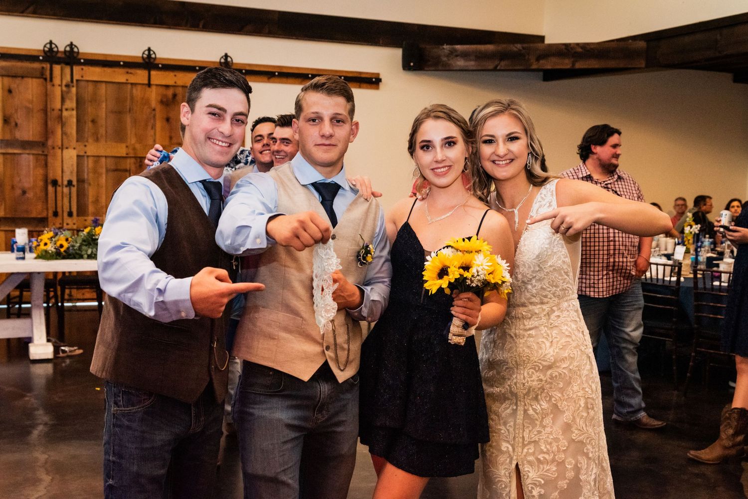 Wedding party poses with drinks and sunflower bouquets at rustic indoor reception.