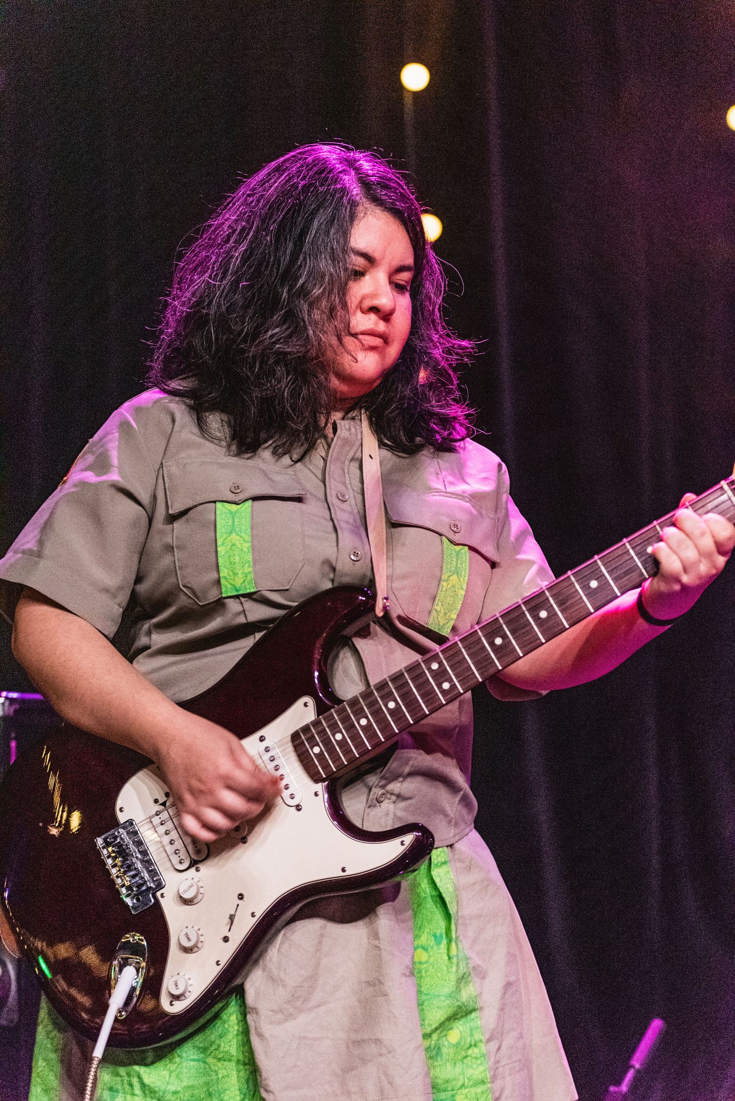 Guitarist in khaki uniform with neon green accents plays Stratocaster guitar during live performance.