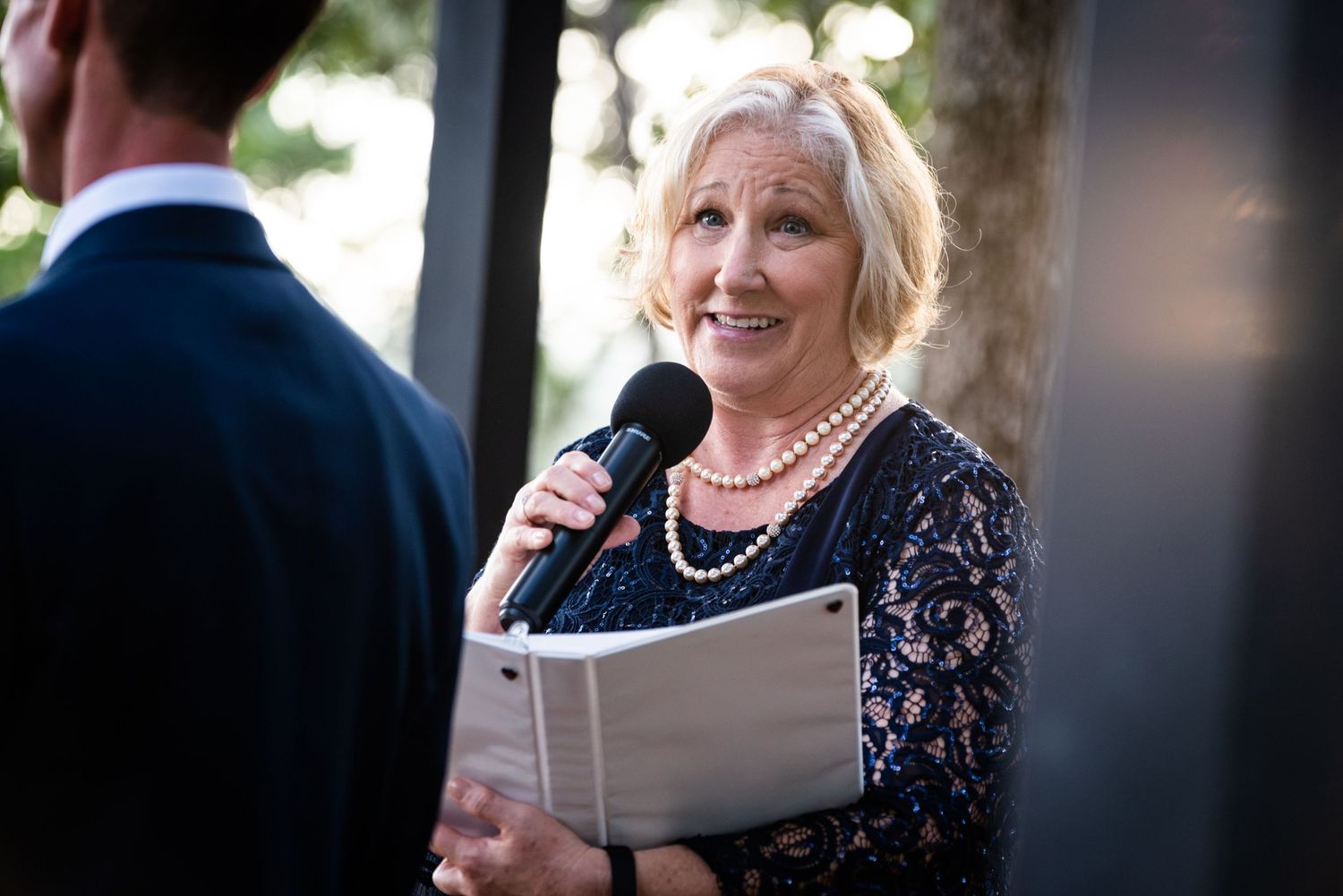 An officiant in navy lace dress holds microphone while reading from ceremony book.