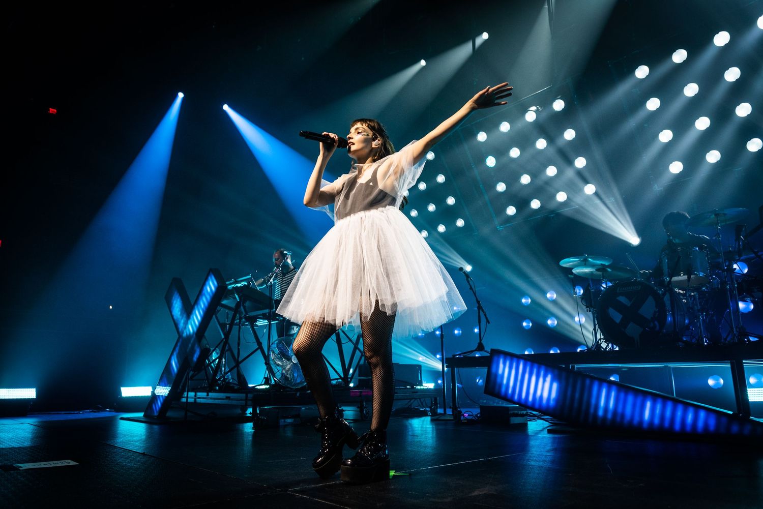 Performers on stage during a concert with dramatic blue lighting and spotlights creating a dynamic atmosphere.