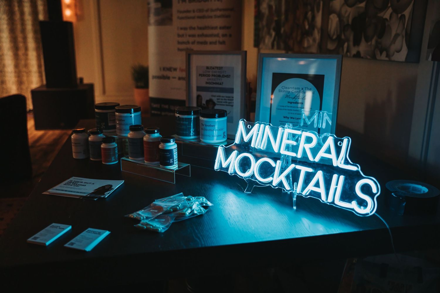 A neon blue sign reading 'MINERAL MOCKTAILS' illuminates a dark bar counter with scattered glassware and items.