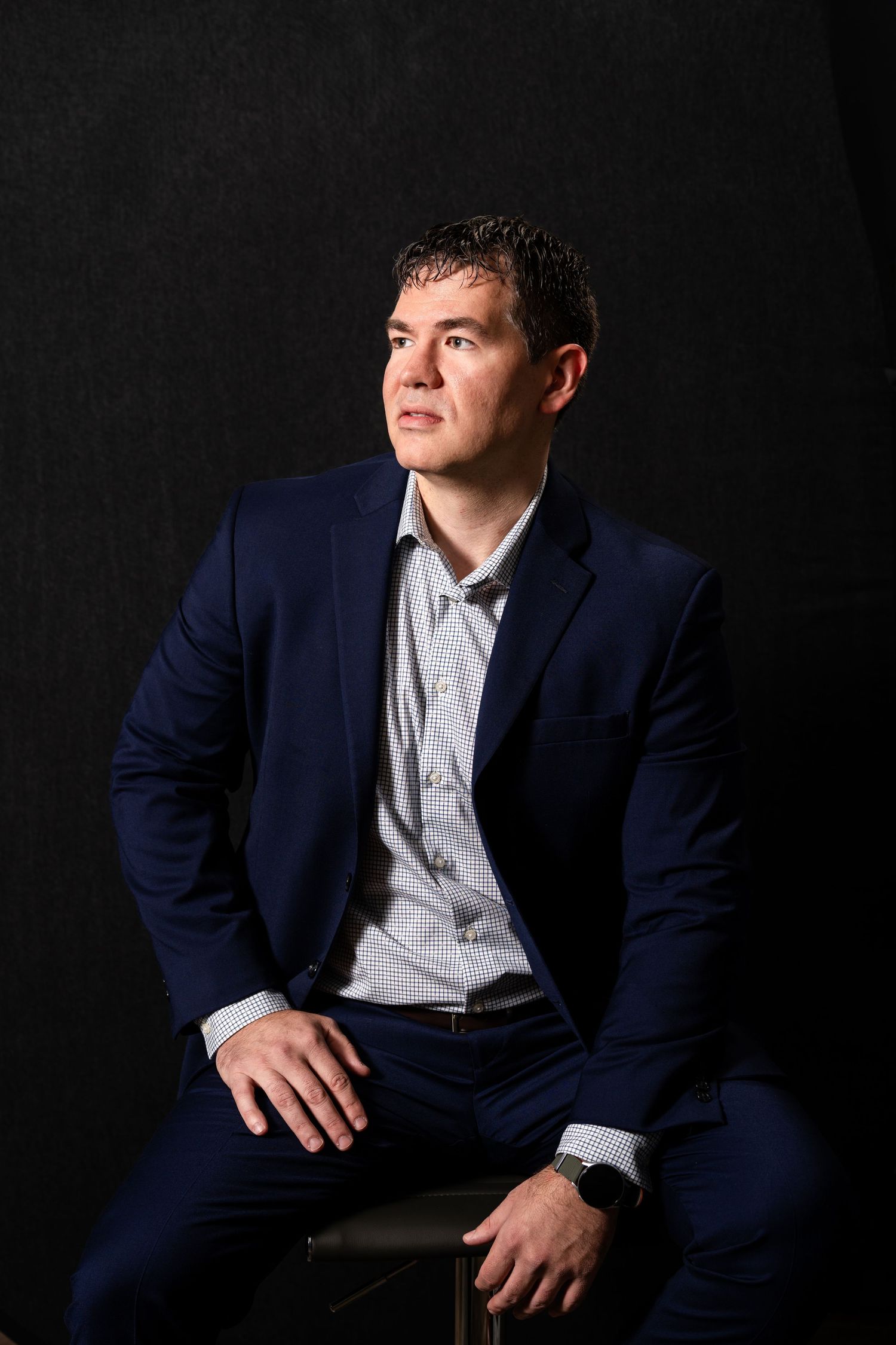 Professional studio portrait of someone in a navy blue suit and light gray dress shirt against a dark background.