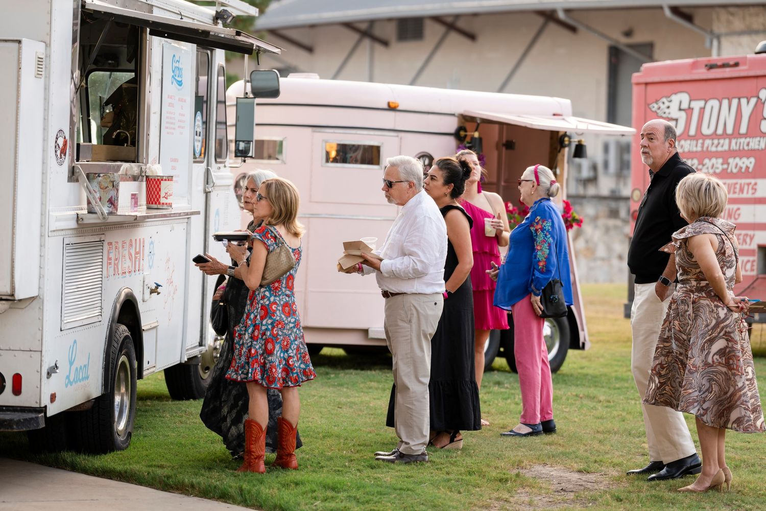 People line up at white food trucks on a grassy area during an outdoor event with colorful summer attire.