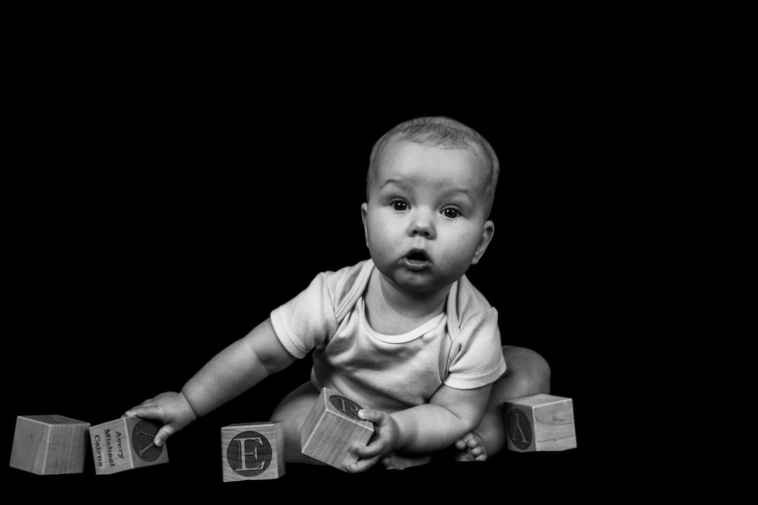 A black and white photo series shows a baby lying on their back holding wooden alphabet blocks.