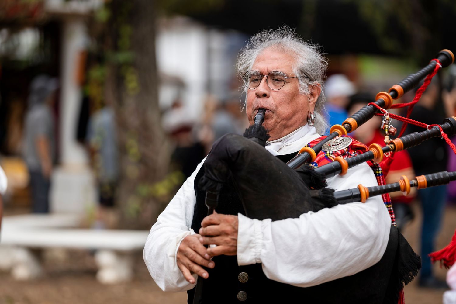 A musician plays traditional bagpipes while wearing a white shirt during an outdoor performance.