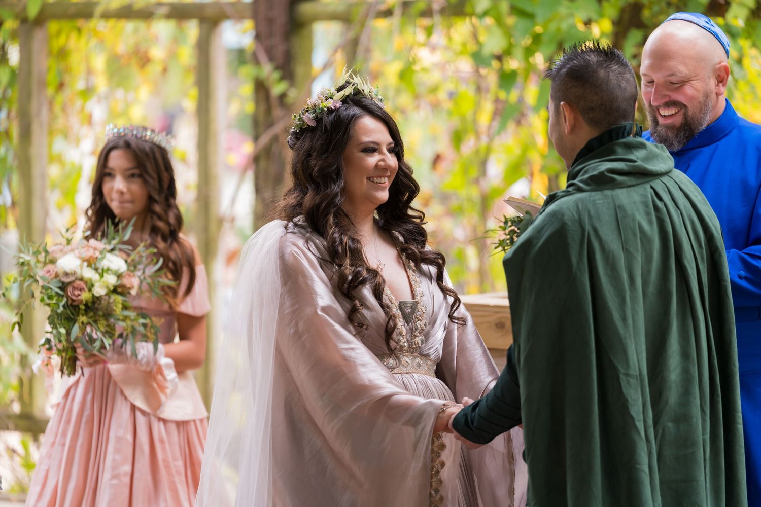 A wedding ceremony takes place outdoors with attendees wearing pink and green attire during a fall day.
