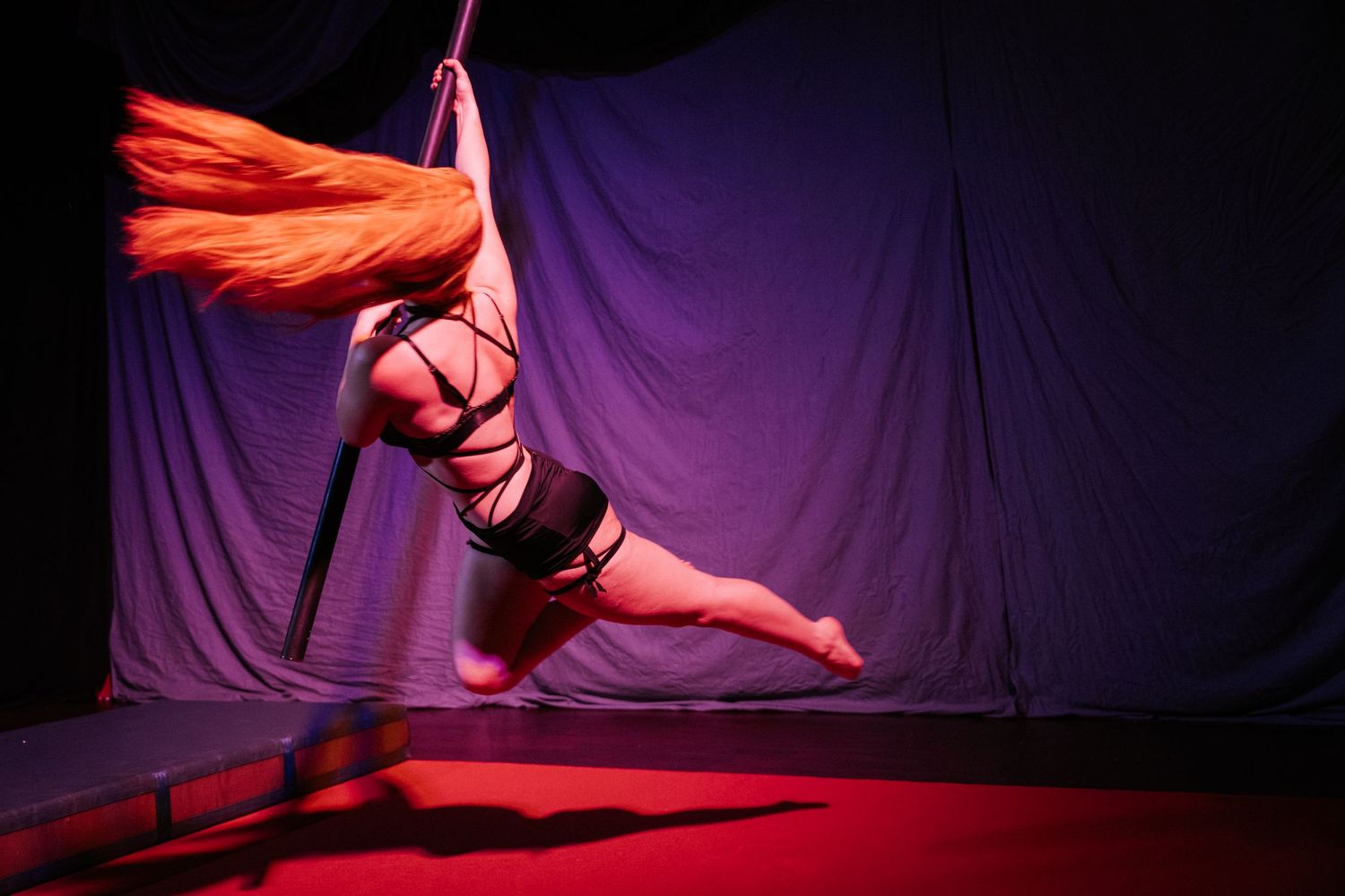 Aerial silk performer suspended mid-air in dramatic red and purple stage lighting against a gray backdrop.