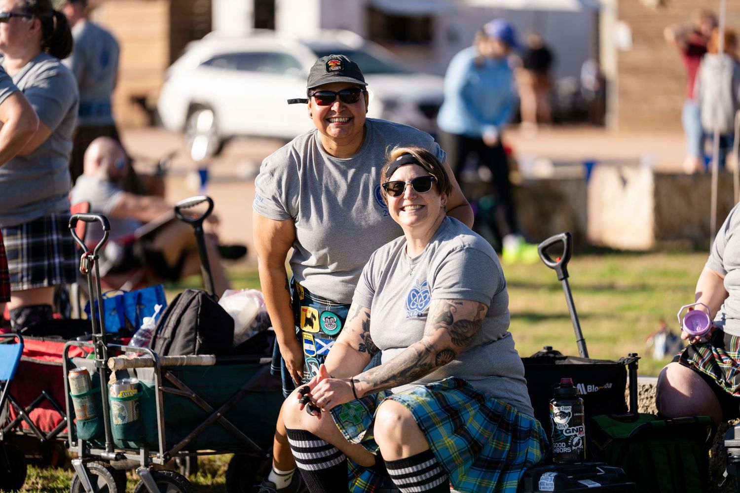 Two people in kilts and casual attire smile at a Scottish Highland Games outdoor event on a sunny day.
