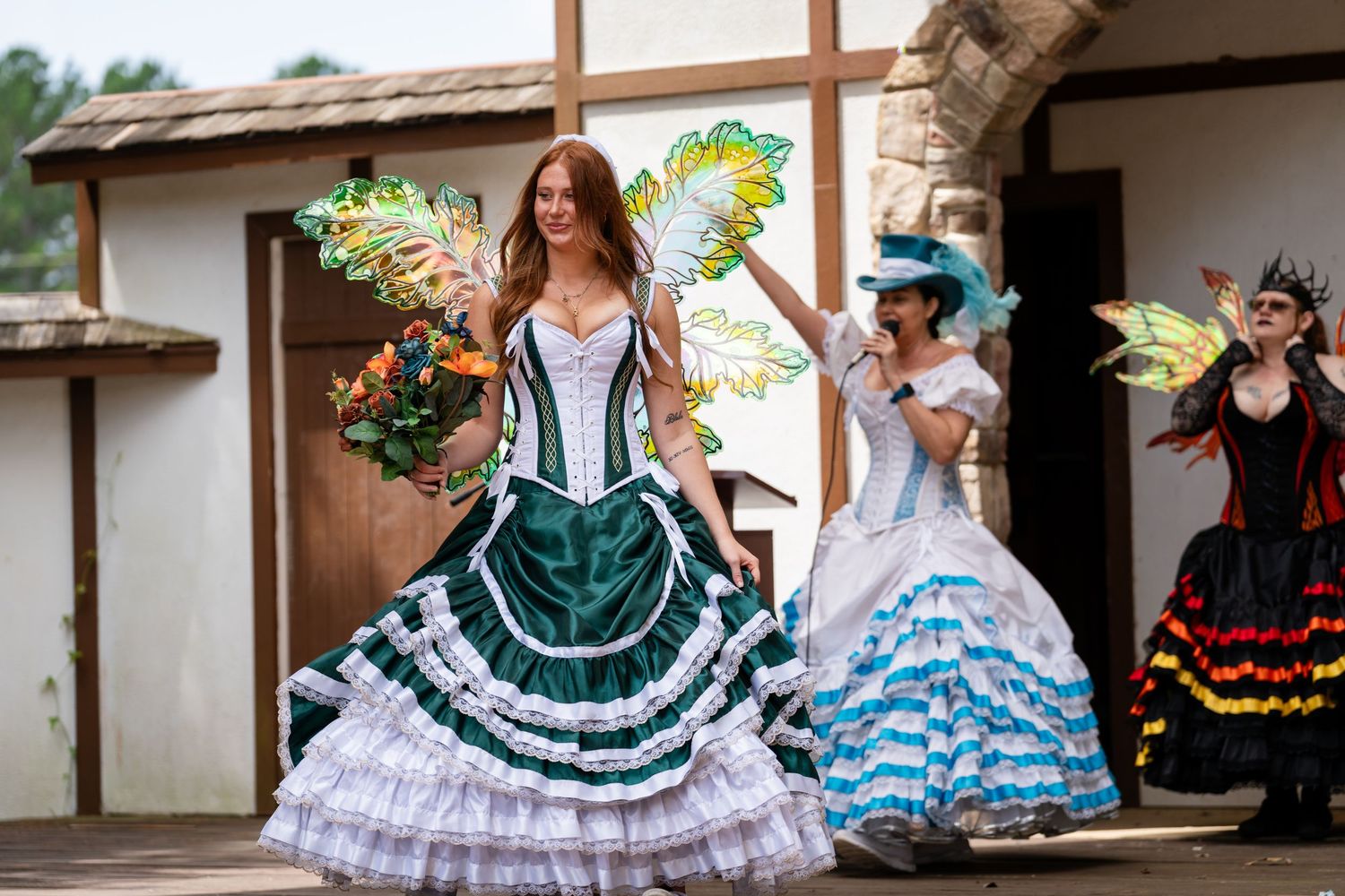 Three women in colorful fairy costumes with wings perform at a Renaissance festival outdoor stage.