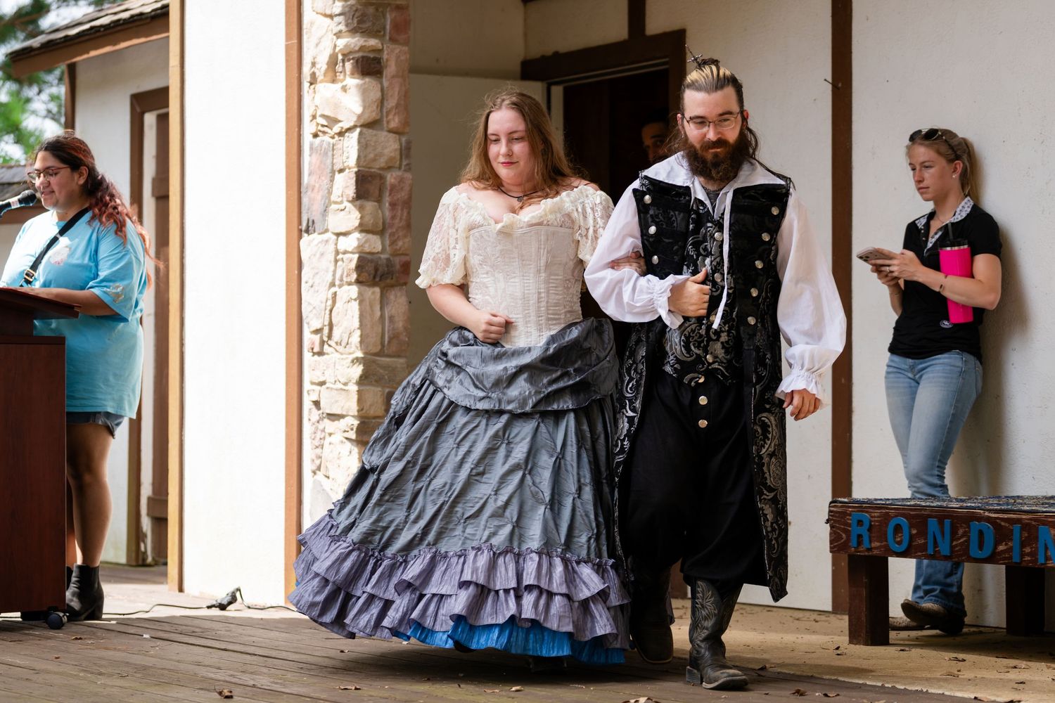Couple in Renaissance faire costumes walk on wooden stage, woman in blue gown, man in black doublet.