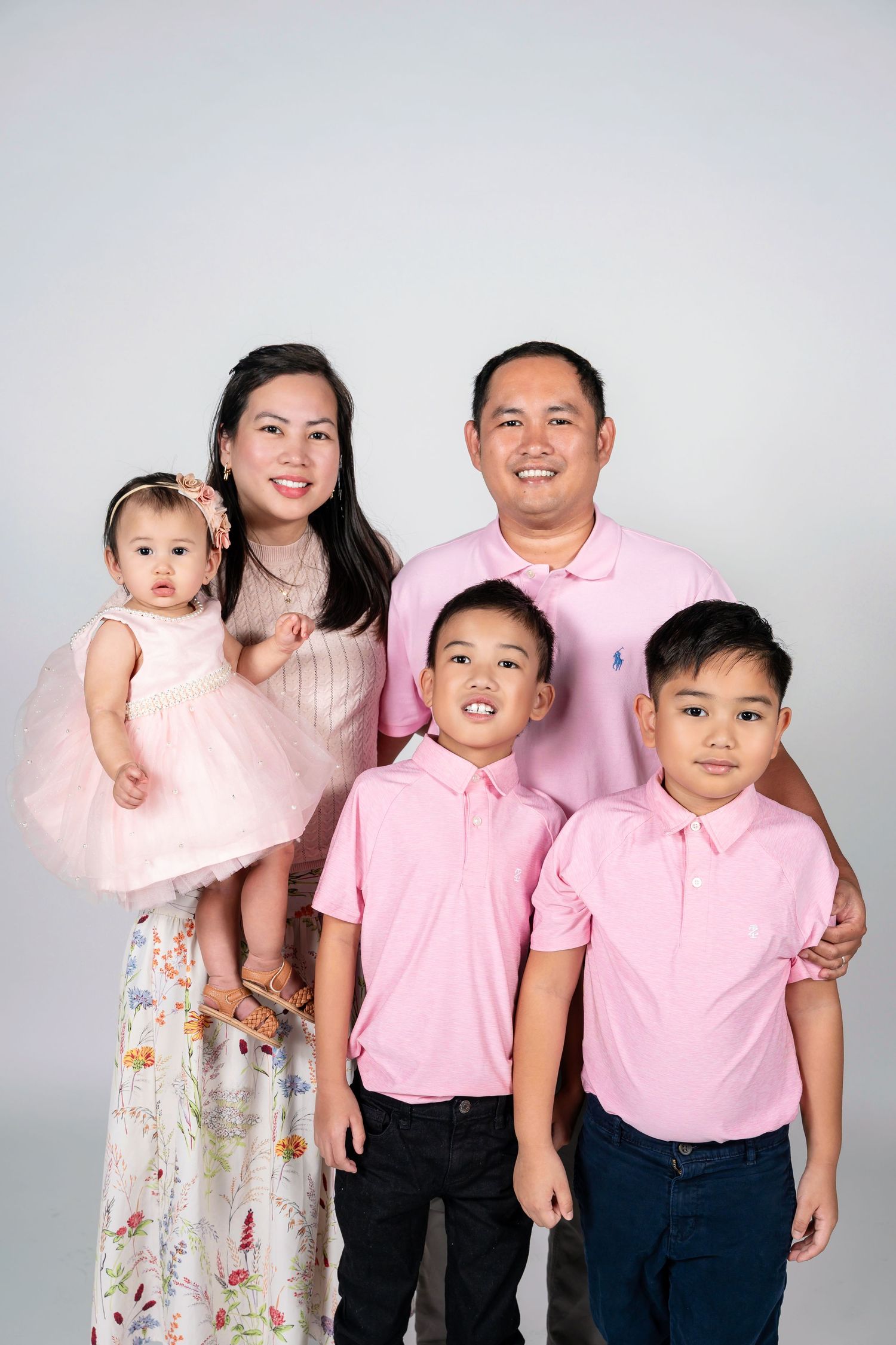 Family of five in matching pink outfits posing for a studio portrait, parents with three young children on white background.