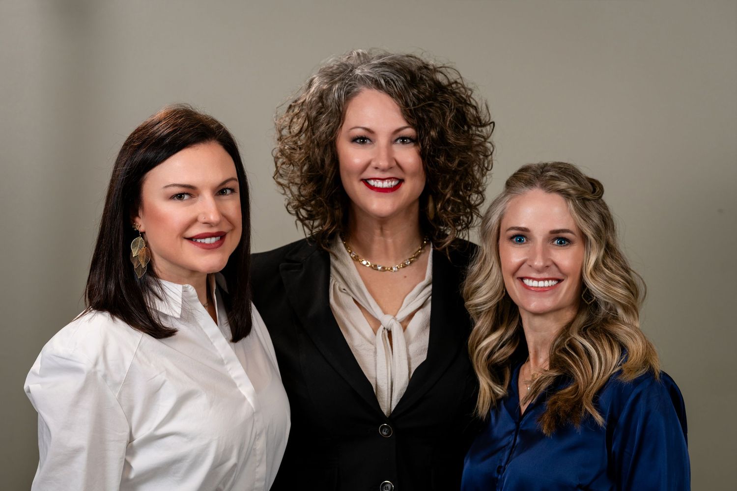 Three professional women smiling together in a formal portrait photo against a neutral gray background.