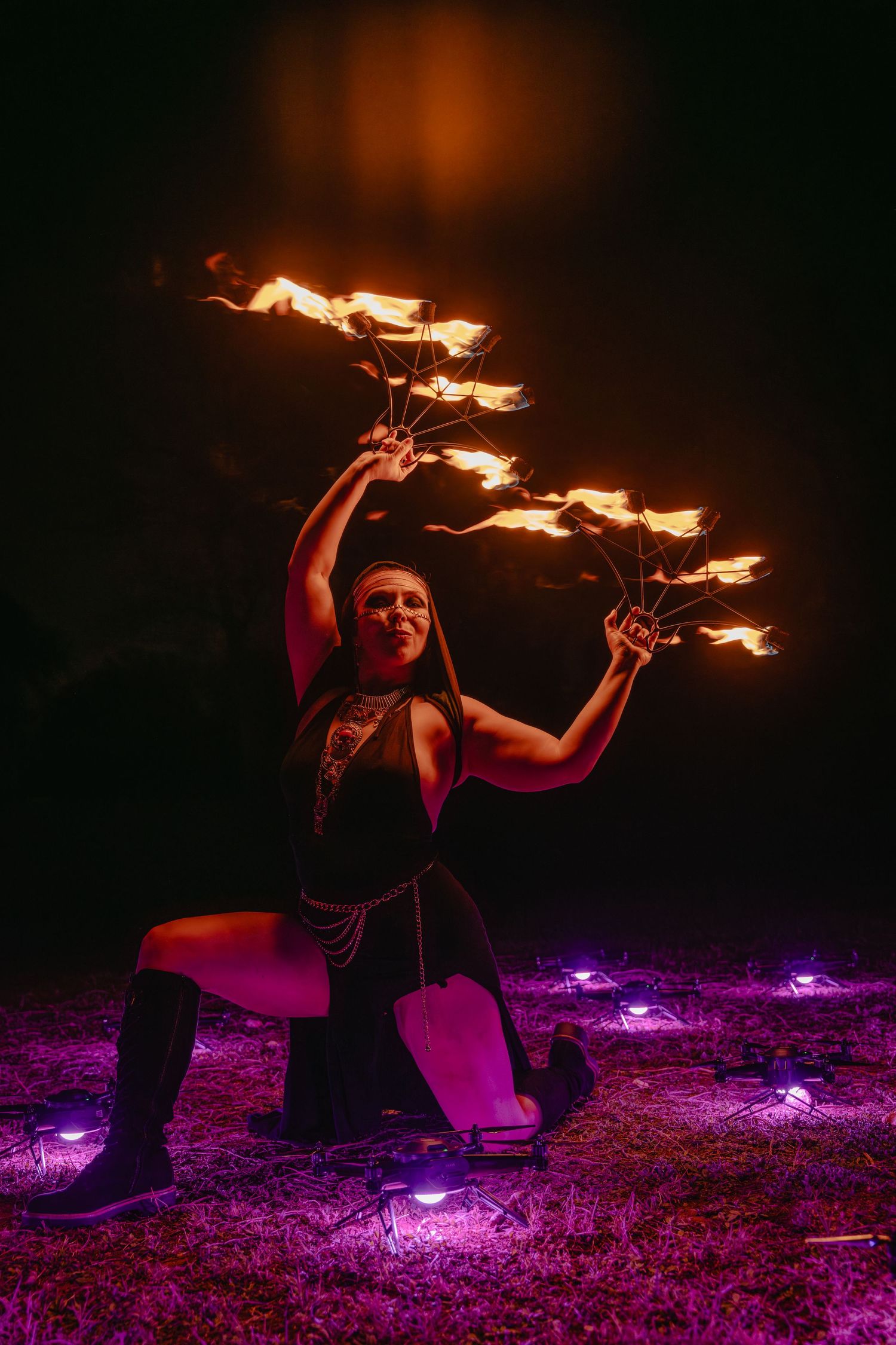 Fire dancer performing at night, holding flaming props aloft, kneeling on grass lit by purple lights.
