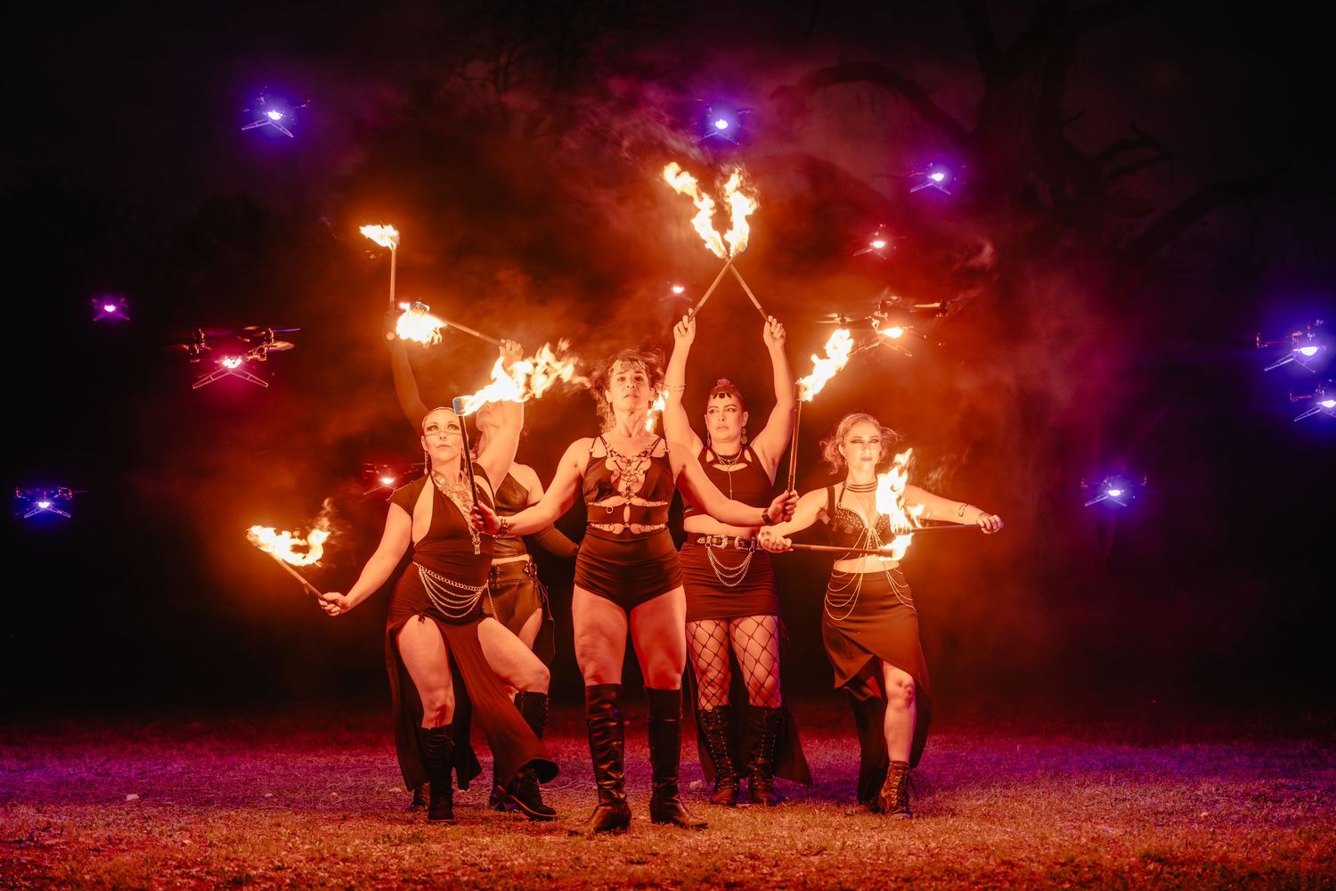 Five performers pose dramatically with fire props against a smoky night sky in a stunning outdoor fire dance show.
