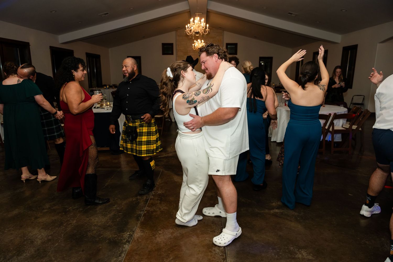 Guests dancing joyfully at a wedding reception in a warmly lit venue with chandelier lighting.