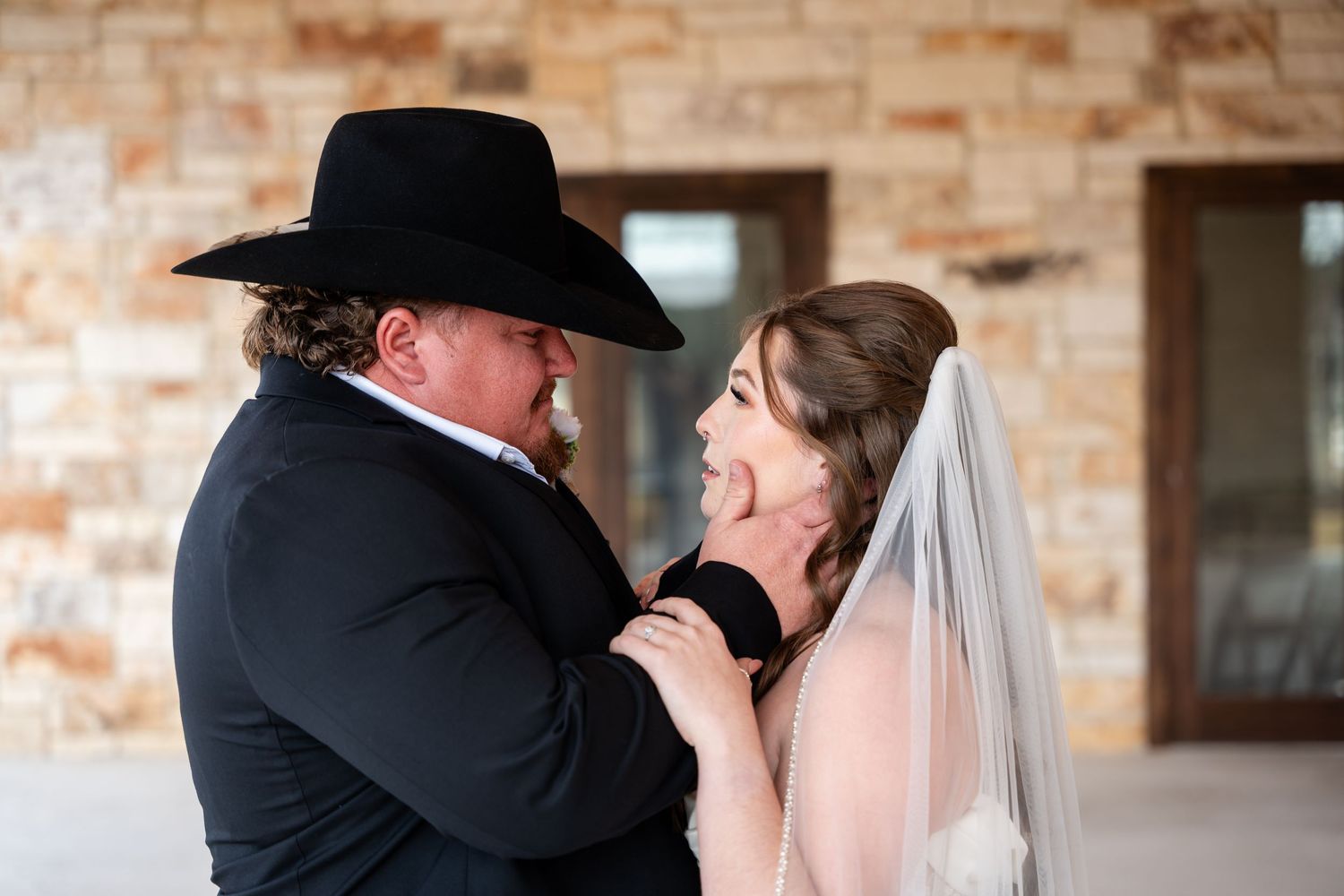 Groom in black cowboy hat tenderly holds bride's face during romantic wedding moment outdoors.