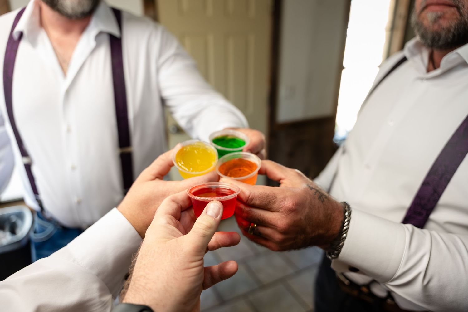 Men in suspenders clinking colorful shot glasses together in a celebratory toast indoors.
