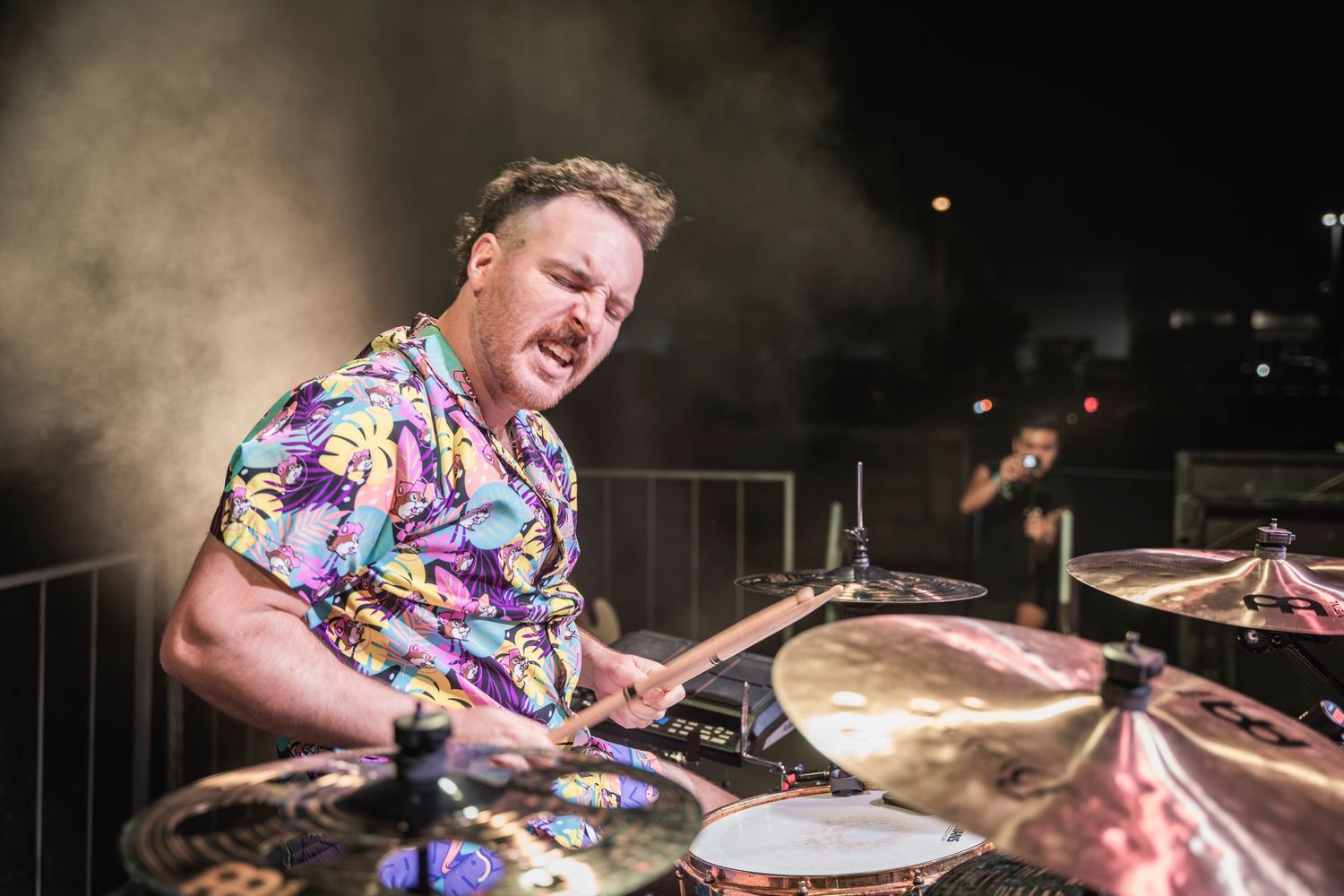 A drummer wearing a colorful Hawaiian shirt performs energetically on a drum kit during a live performance.