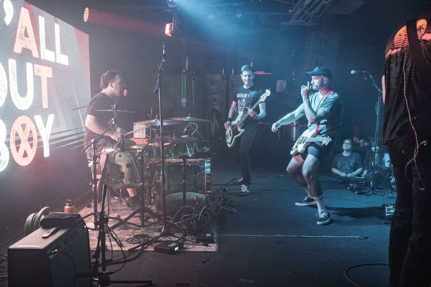 Pop punk band performs energetically on a dark stage with blue lighting and 'All Out' text visible in background.