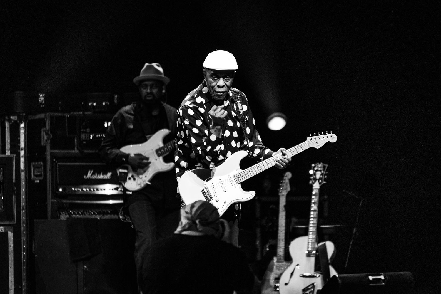 Black and white photo of musicians performing on stage with guitars and amplifiers at a live concert.