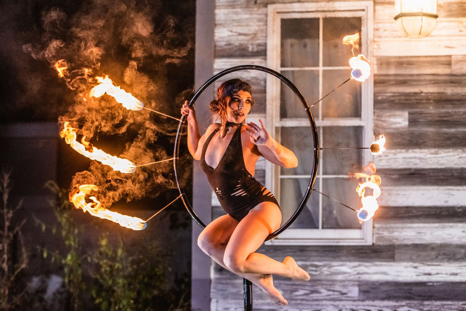 Fire performer spins flaming batons in a dramatic dance sequence against a rustic wooden building backdrop.