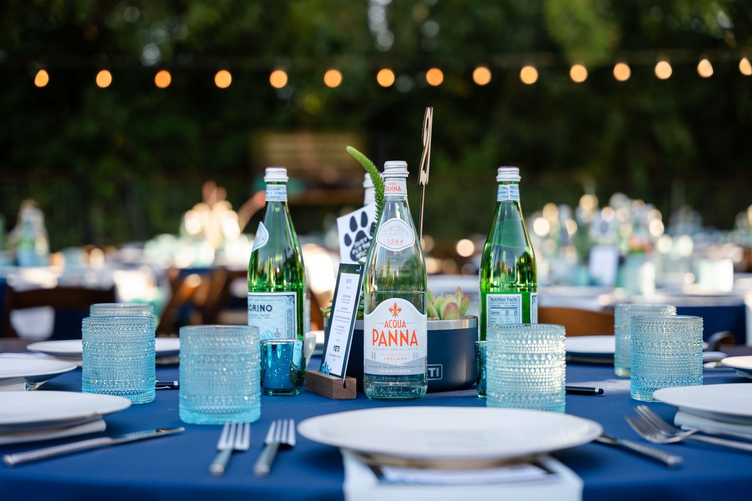 Wedding reception table settings with blue glass bottles and string lights creating an elegant outdoor ambiance.