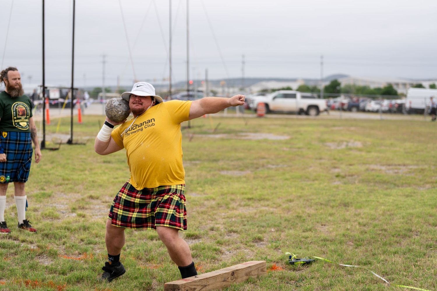 Man in yellow shirt and tartan kilt throws shot put at Highland Games competition outdoors.