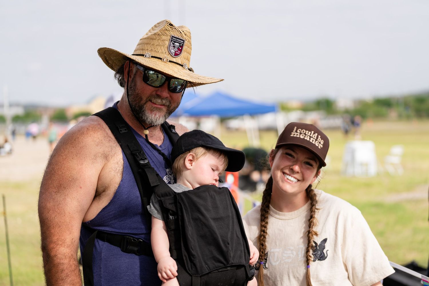 Family of three at outdoor festival, father with straw hat carrying baby in carrier beside smiling woman with braids.