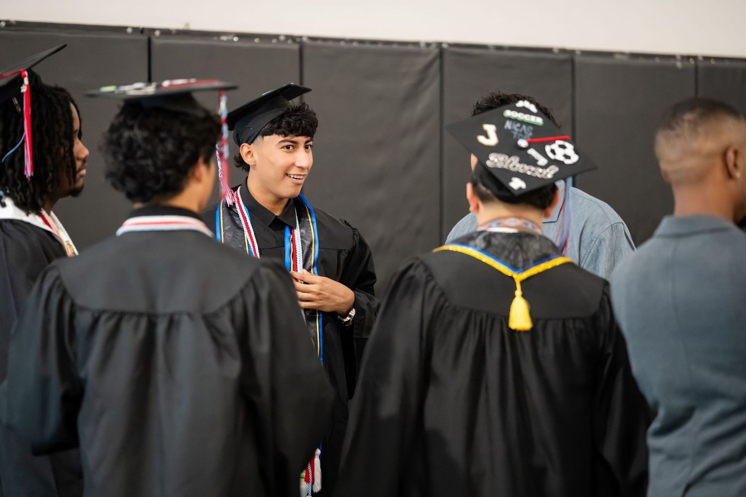 Graduates in black caps and gowns socializing before a graduation ceremony in a school gymnasium.