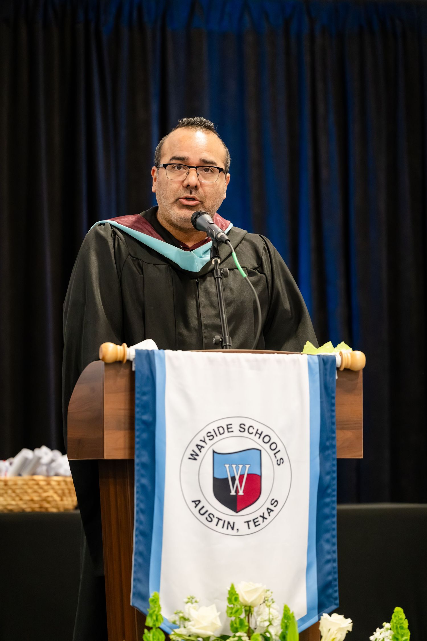 Man in graduation regalia speaks at Wayside Schools Austin Texas podium during commencement ceremony.